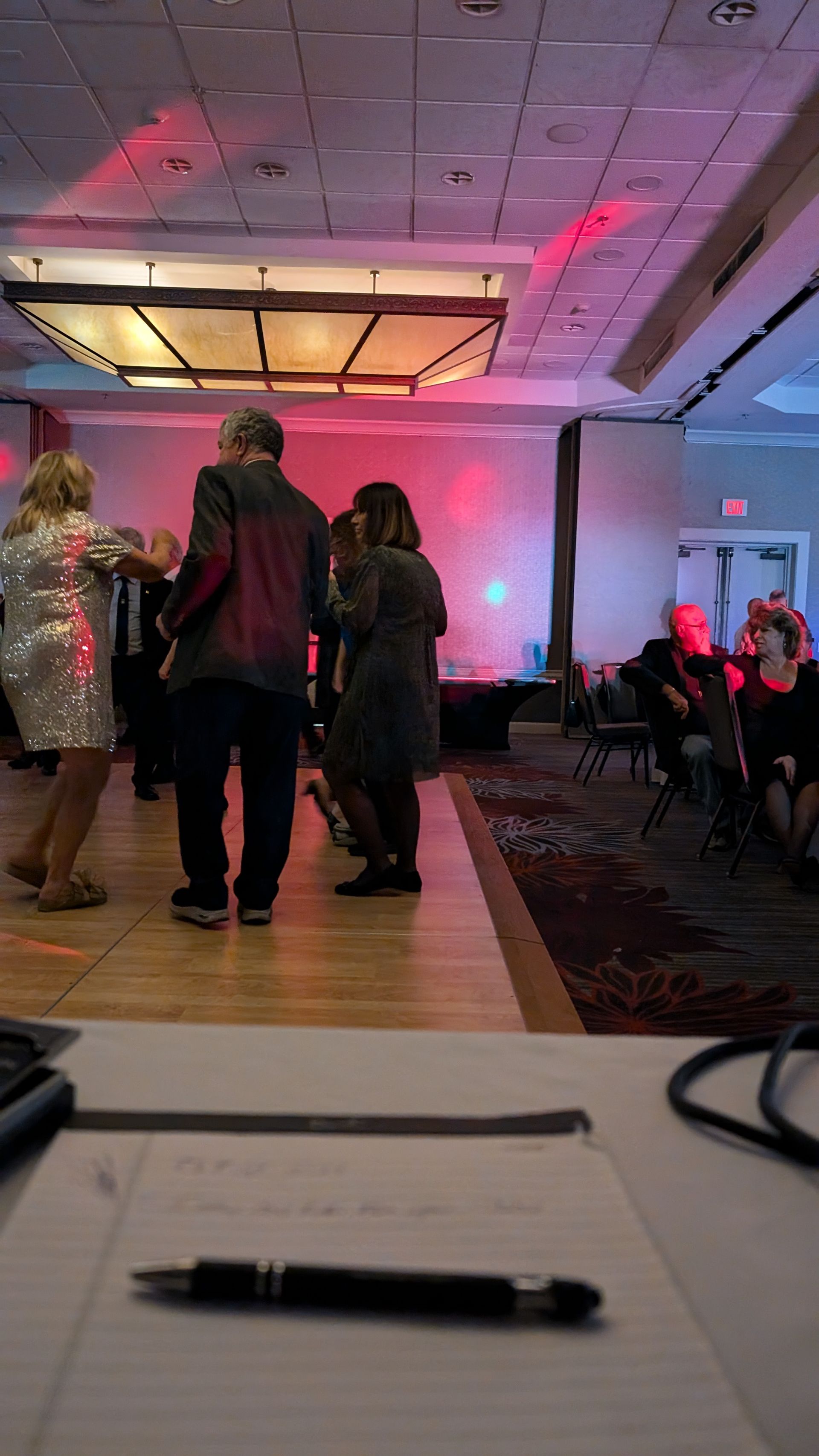 People dancing on a lit banquet hall dance floor with seated guests nearby