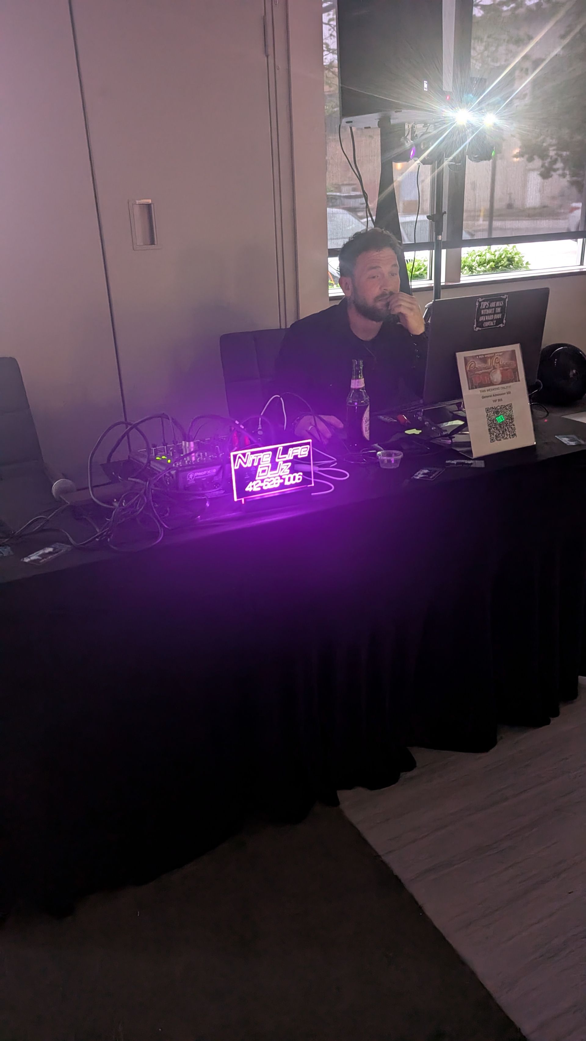 Person seated behind a black table with purple-lit audio equipment and a display card at an indoor event.