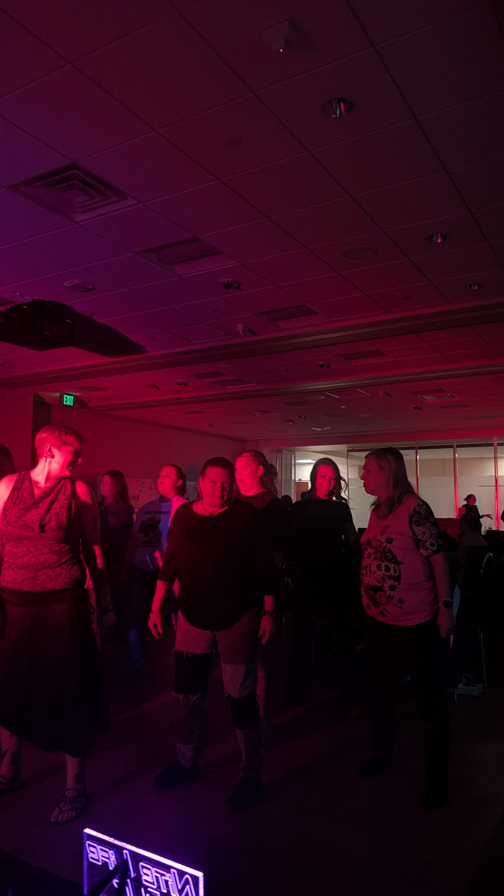 Small group of people standing in a dim room under pink and red lighting, with a glowing sign in the foreground
