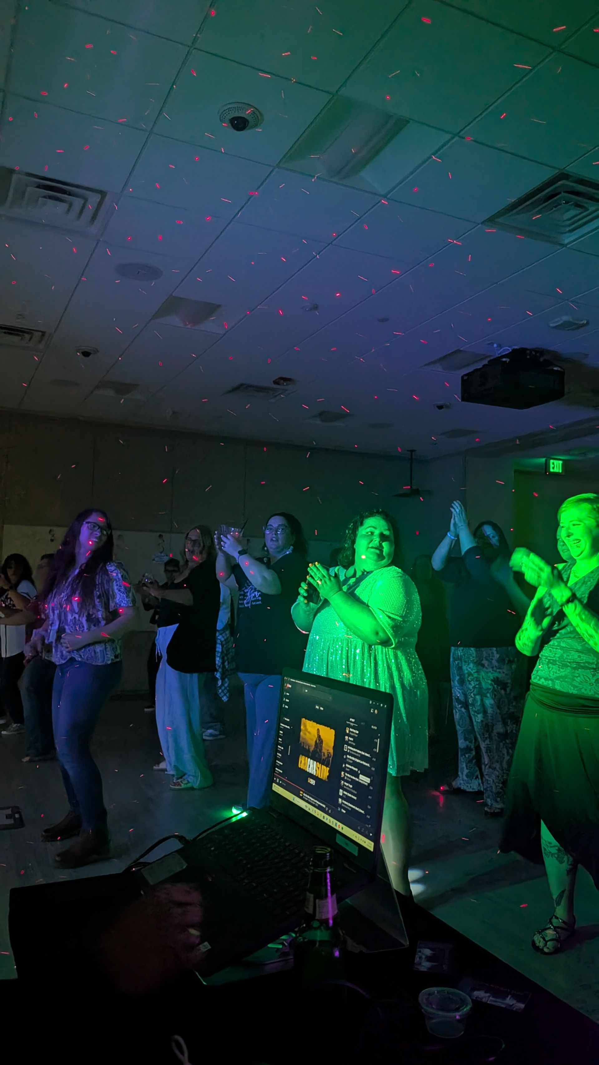 People dancing in a dimly lit room with colorful green and purple stage lights.