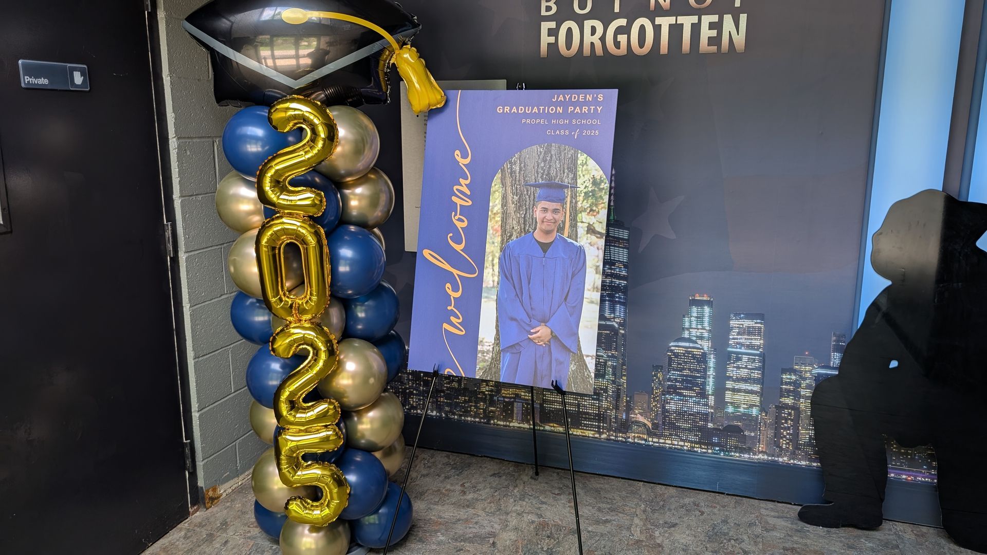 Graduation-themed memorial display with gold and black balloons, portrait banner, and city skyline backdrop