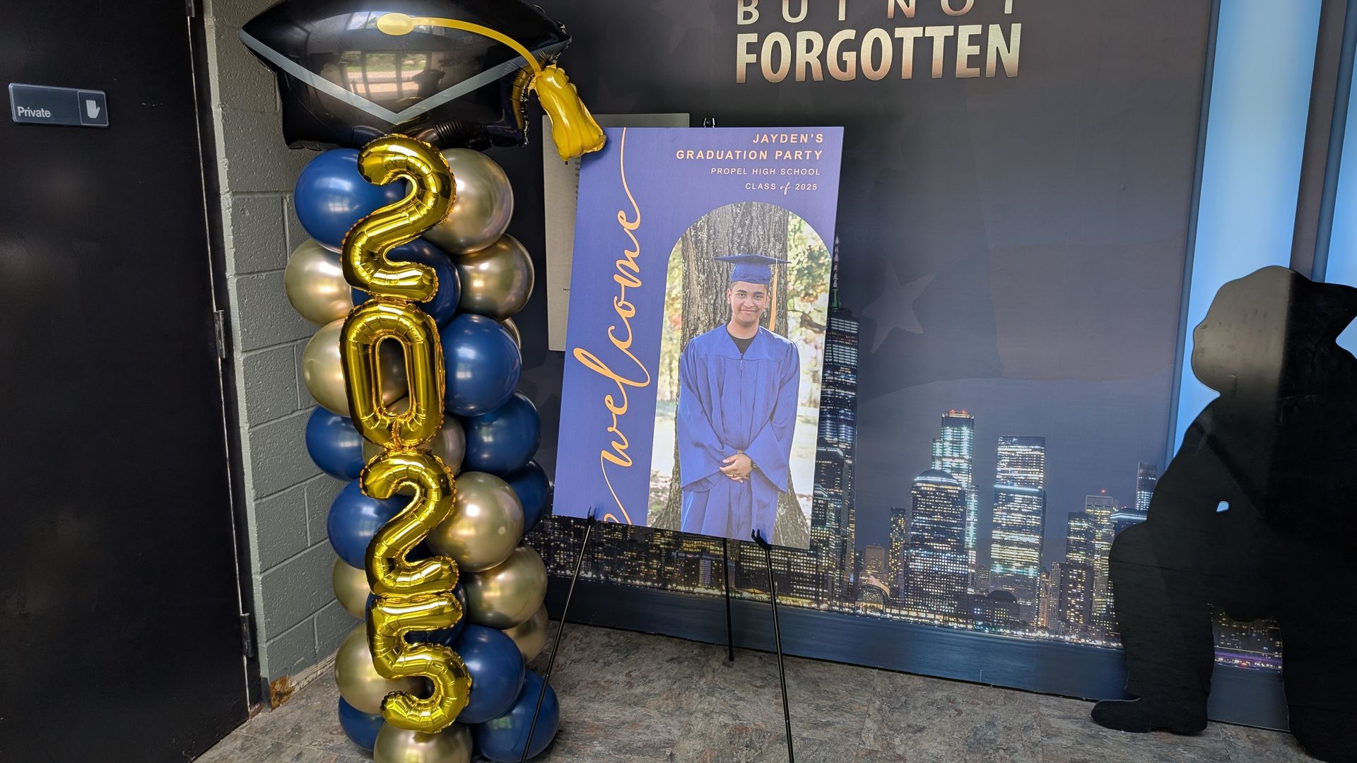 Memorial display with gold balloons, a framed tribute photo, and city skyline backdrop on a dark table