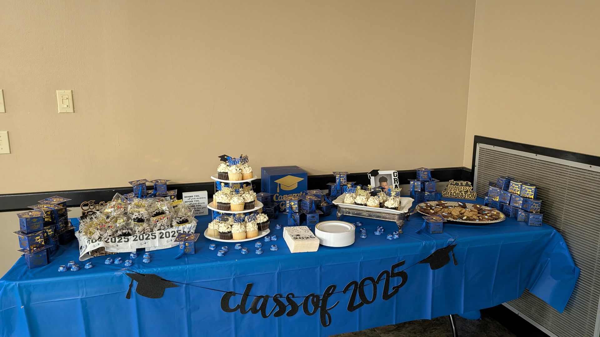 Dessert table with blue tablecloth reading “Class of 2025,” covered in snacks and treats.