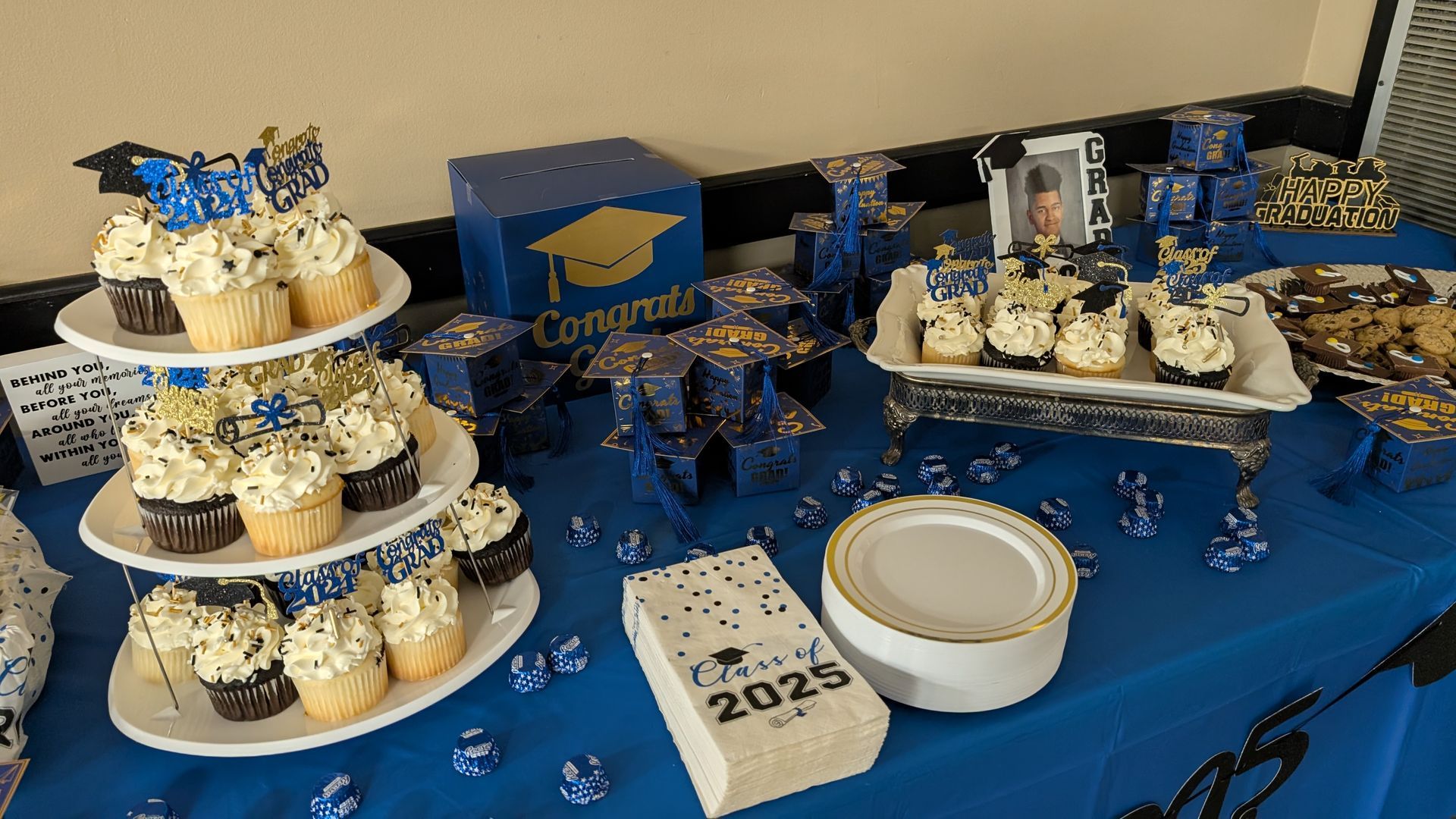 Blue graduation dessert table with cupcakes, cakes, and gold accents, including a “Congrats” sign and 2025 decoration