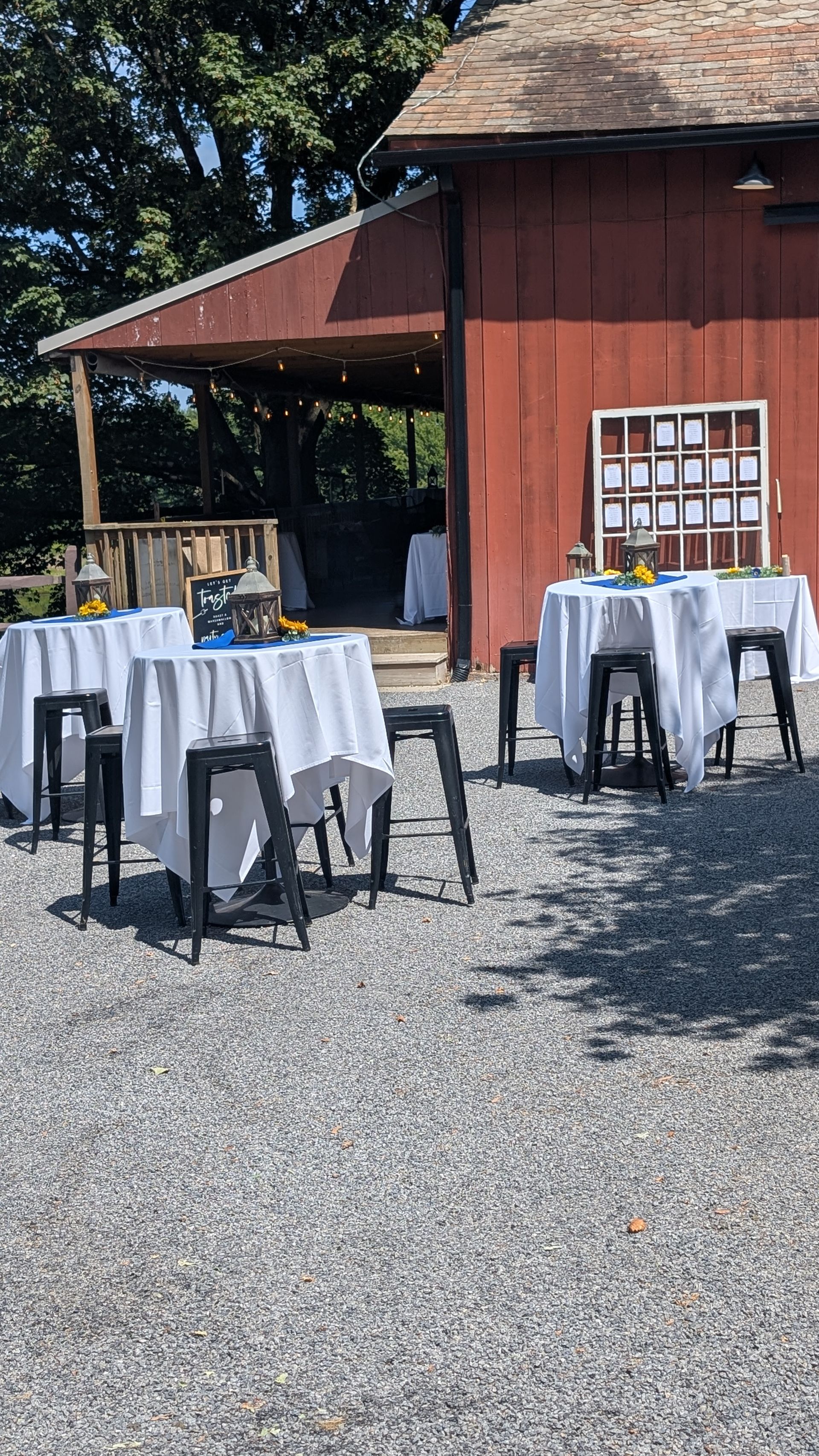 Outdoor patio with round tables, white tablecloths, black stools, and a red barn-like building in sunlight