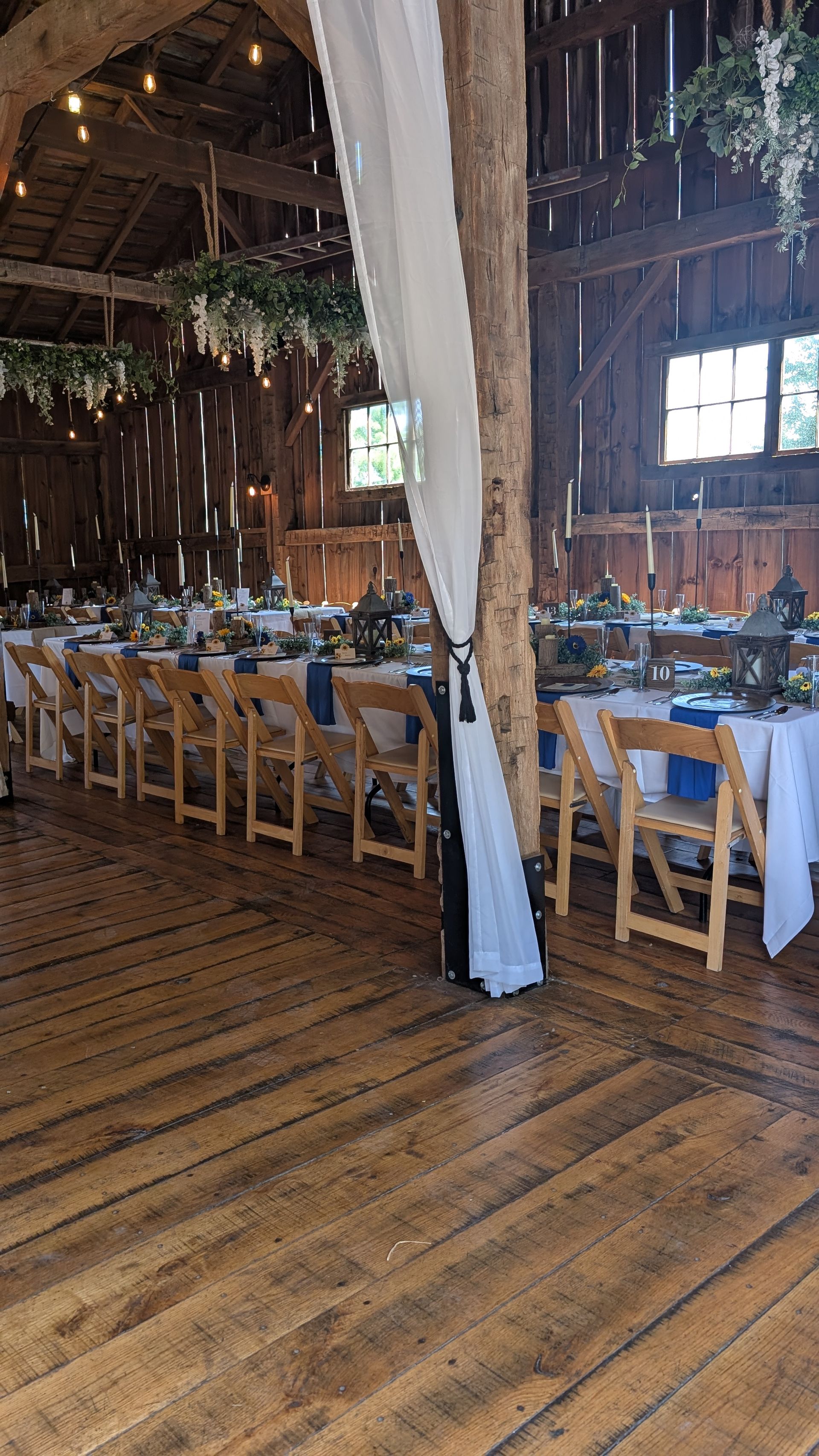 Rustic barn-style dining hall with long tables, wooden chairs, and hanging greenery under a high ceiling