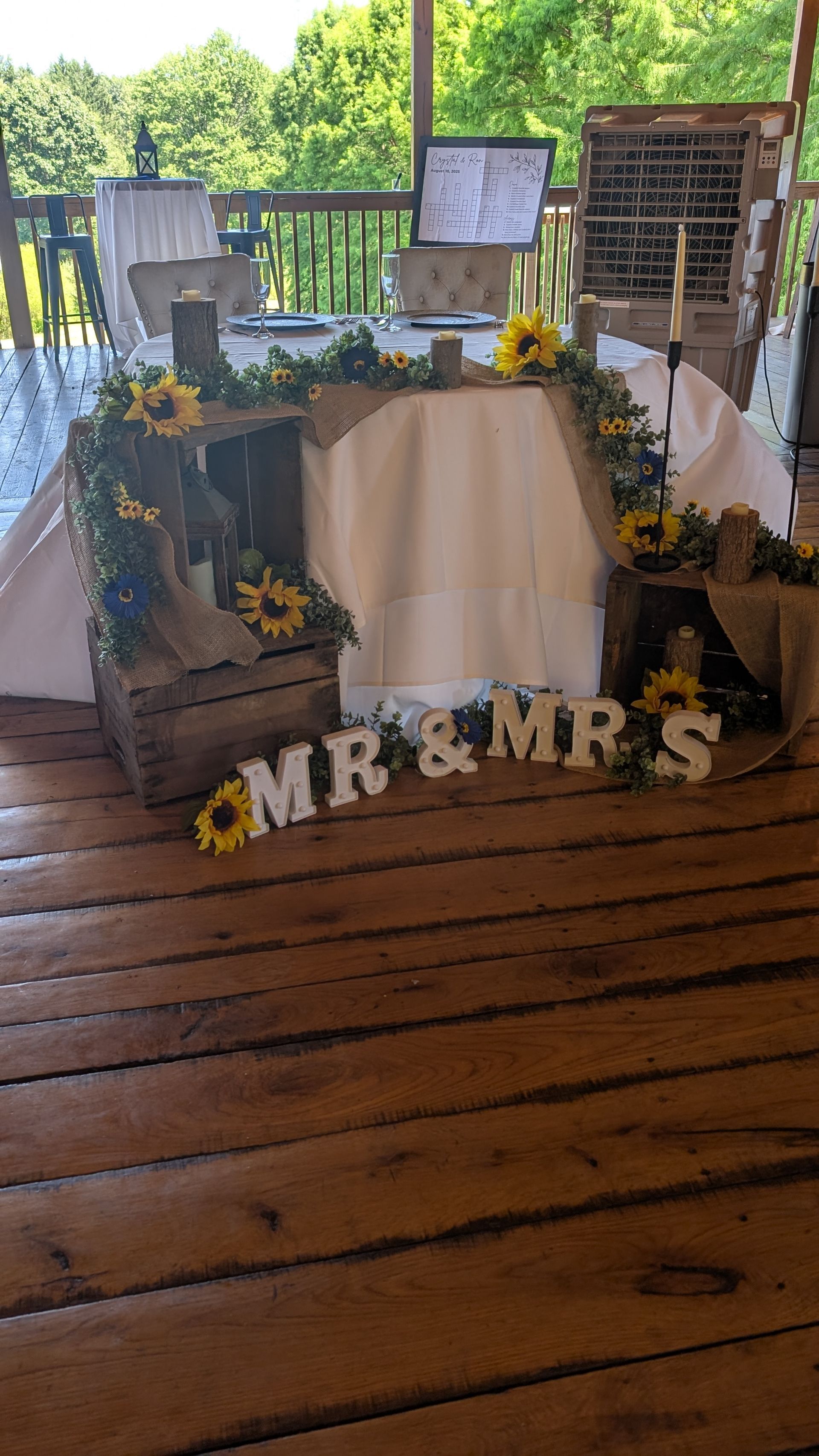 Outdoor decorated table with white cloth, sunflowers, framed photos, and “MR & MRS” letters on a wooden deck