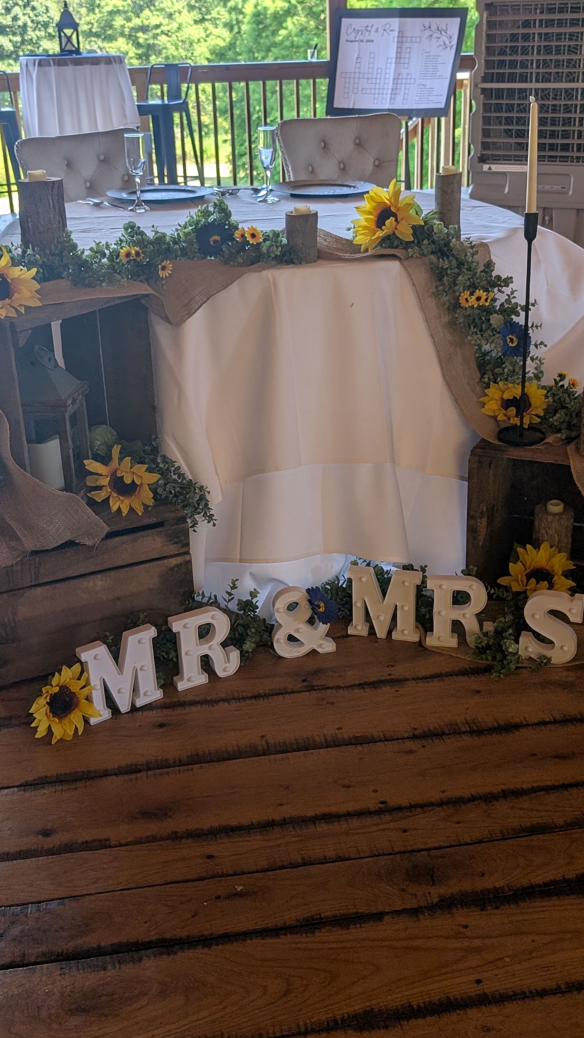 Wedding sweetheart table with white drape, sunflowers, and “MR & MRS” letters on a wooden floor.