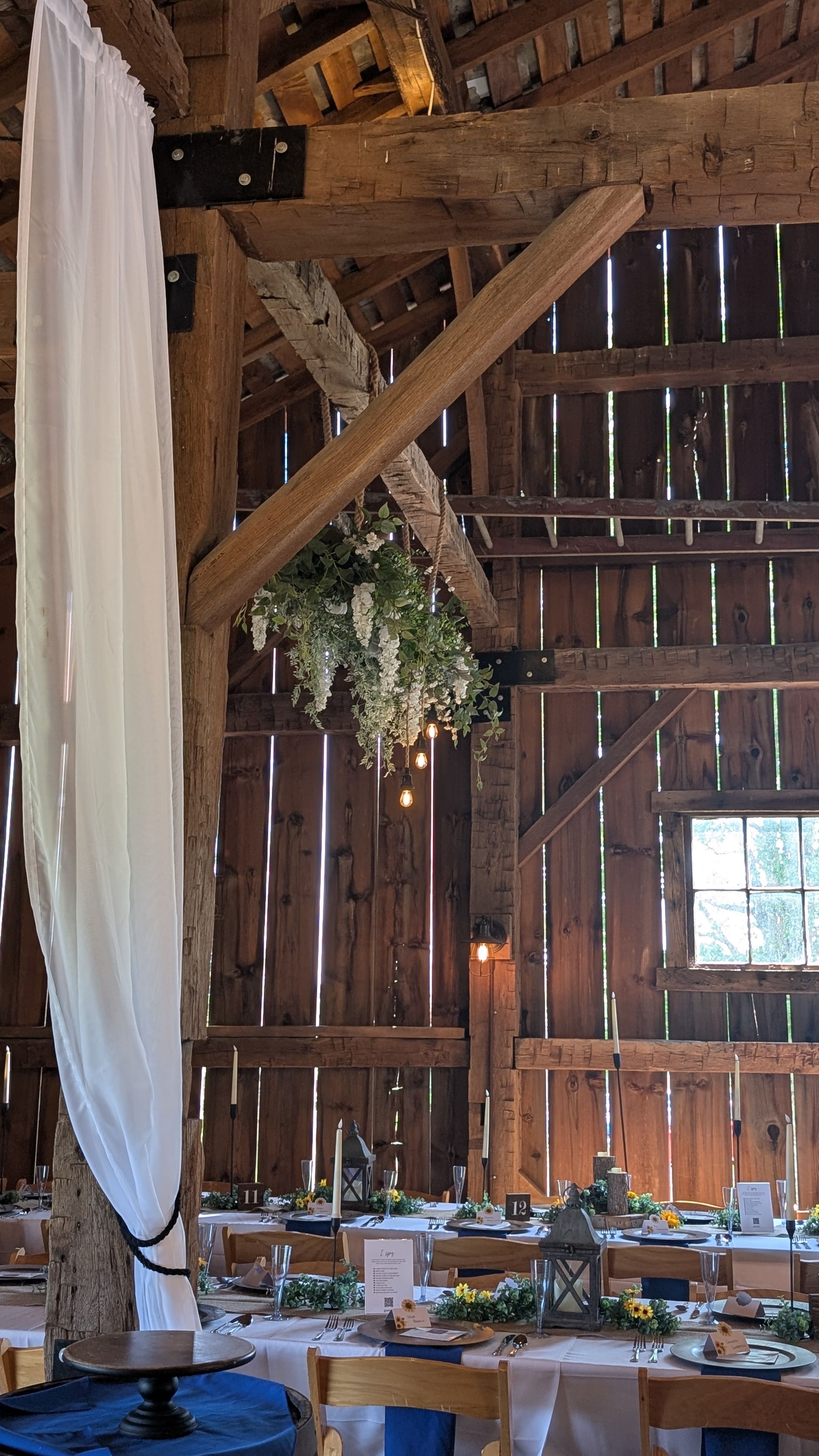 Rustic barn interior with long banquet tables set for an event, hanging greenery, and white drapery.
