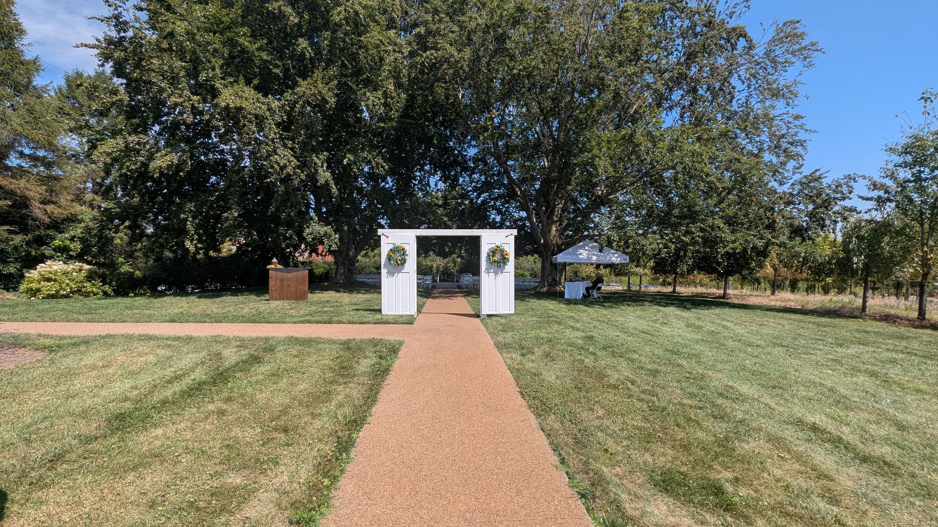 Gravel path leading to a white archway in a grassy park, framed by trees under a clear blue sky
