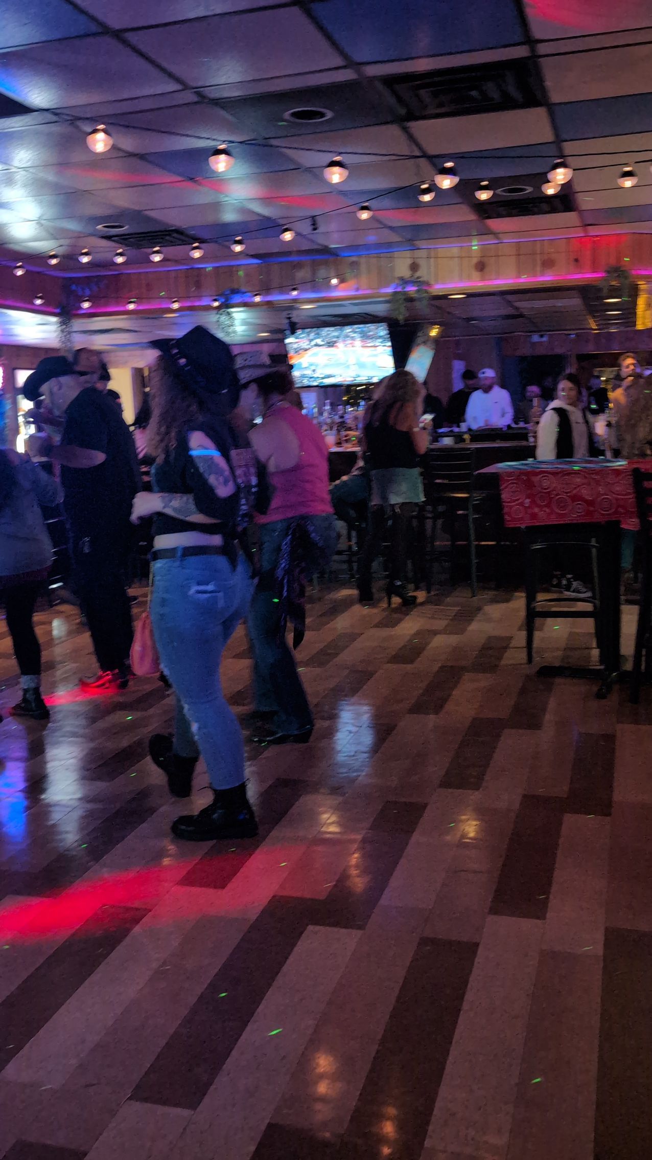 People dancing in a dimly lit nightclub with colorful lights and tables nearby