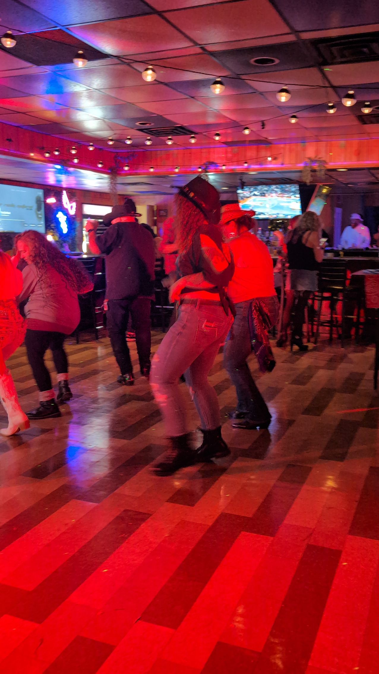 People dancing in a red-lit nightclub with a checkered floor and TVs in the background