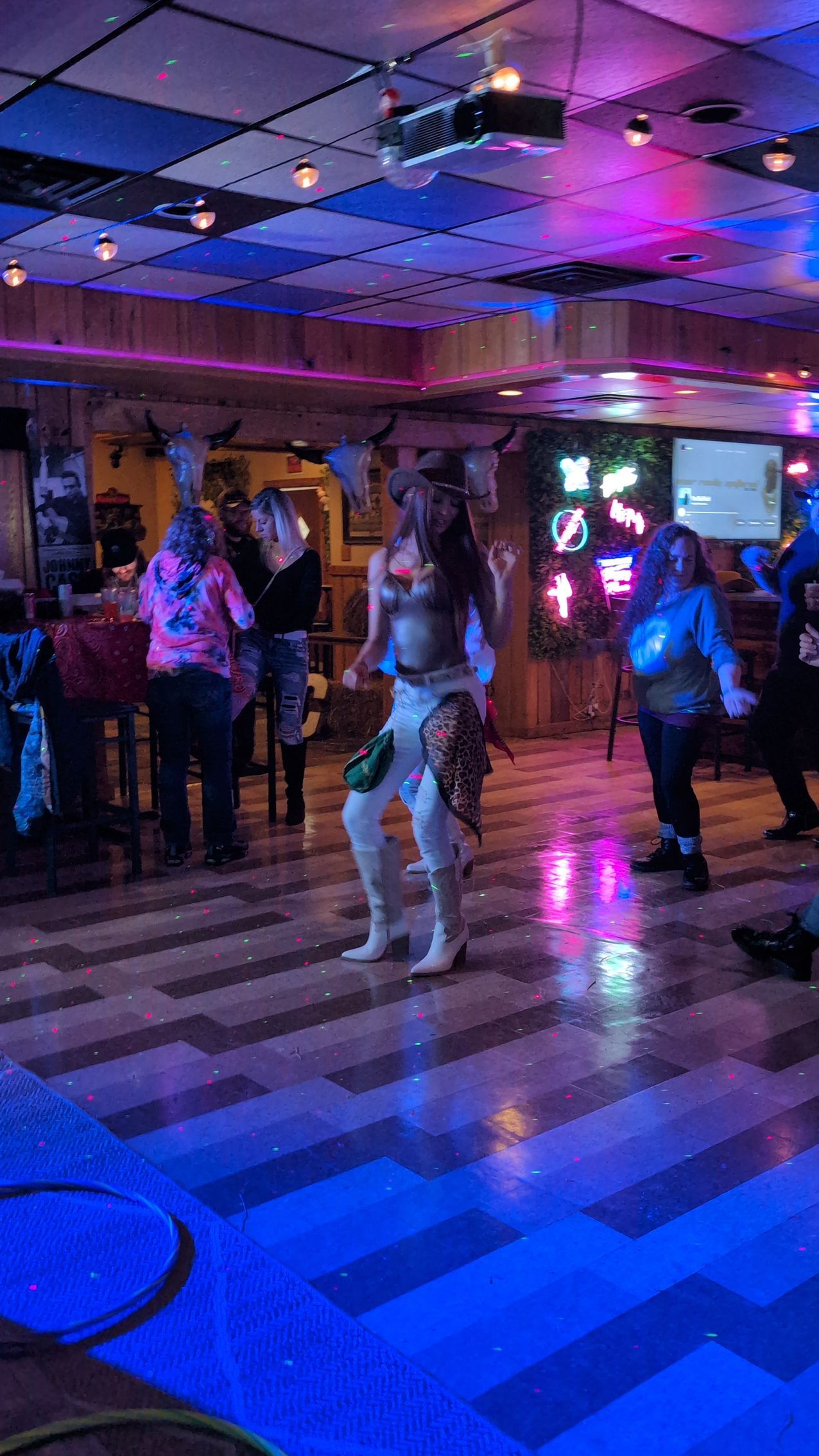 People dancing in a neon-lit hall with reflective floor and purple-blue lighting