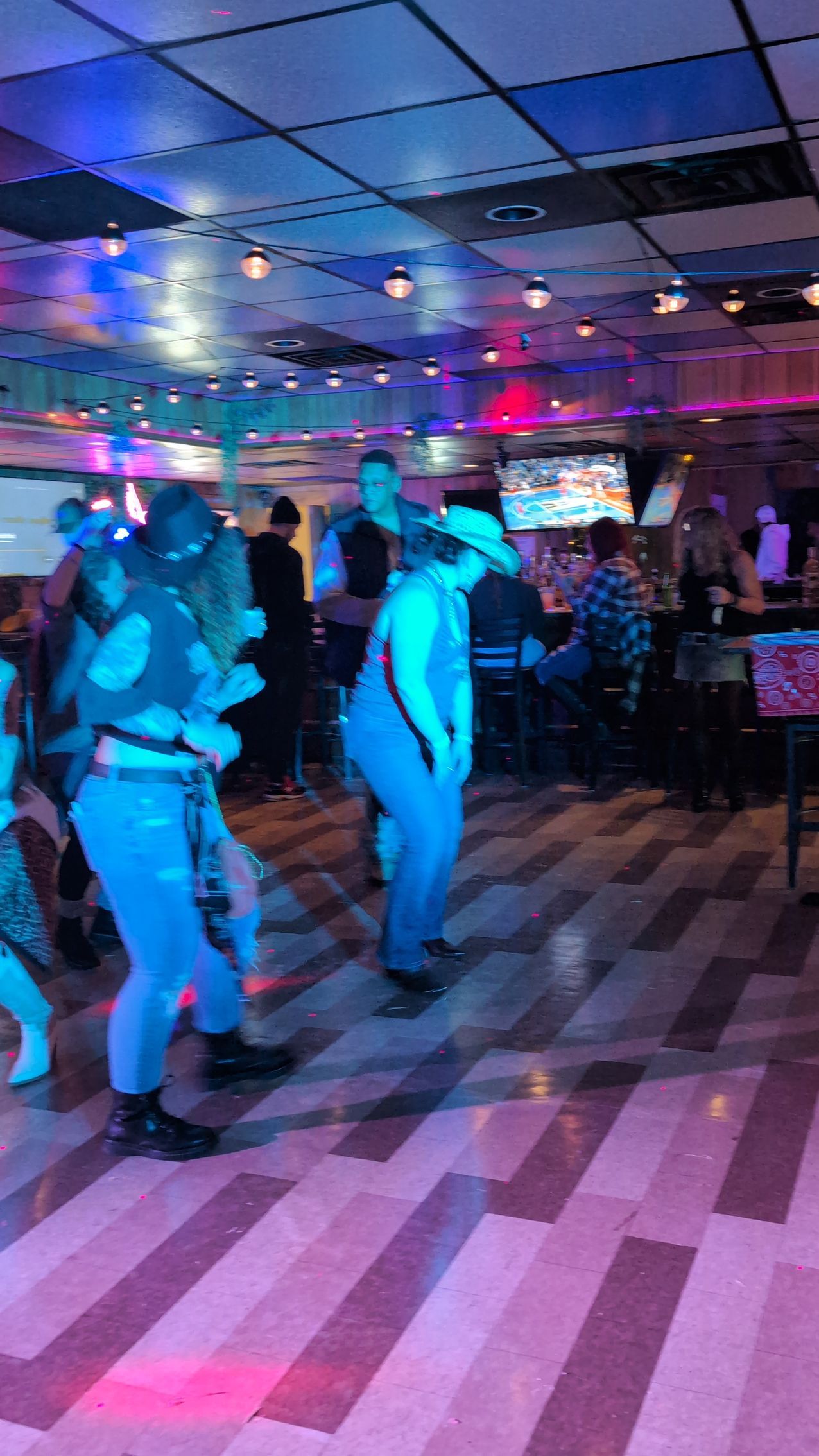 People dancing in a neon-lit club with a checkered floor and colorful ceiling lights.