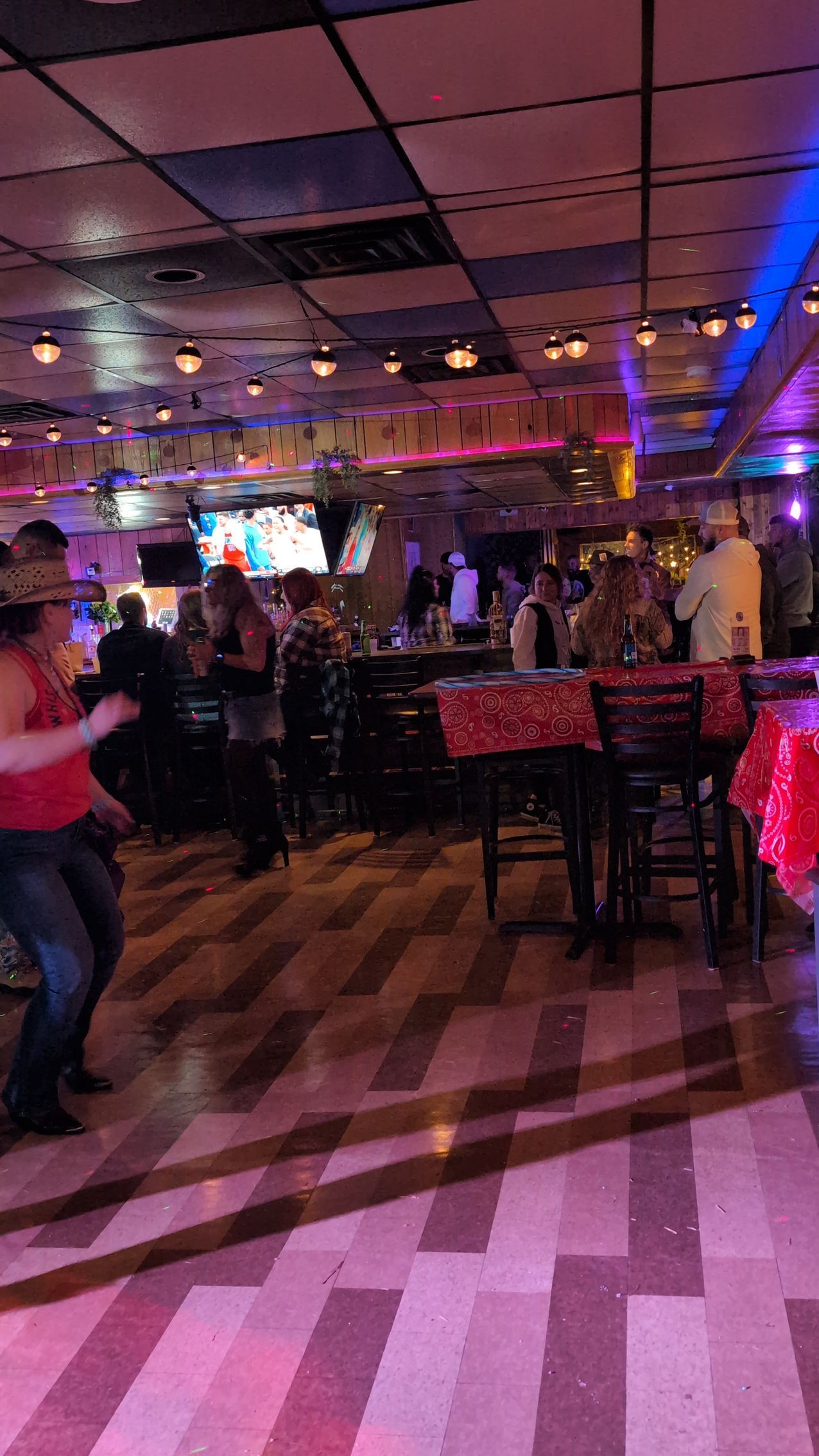Dimly lit bar with people socializing, colorful lights, and checkered floor.