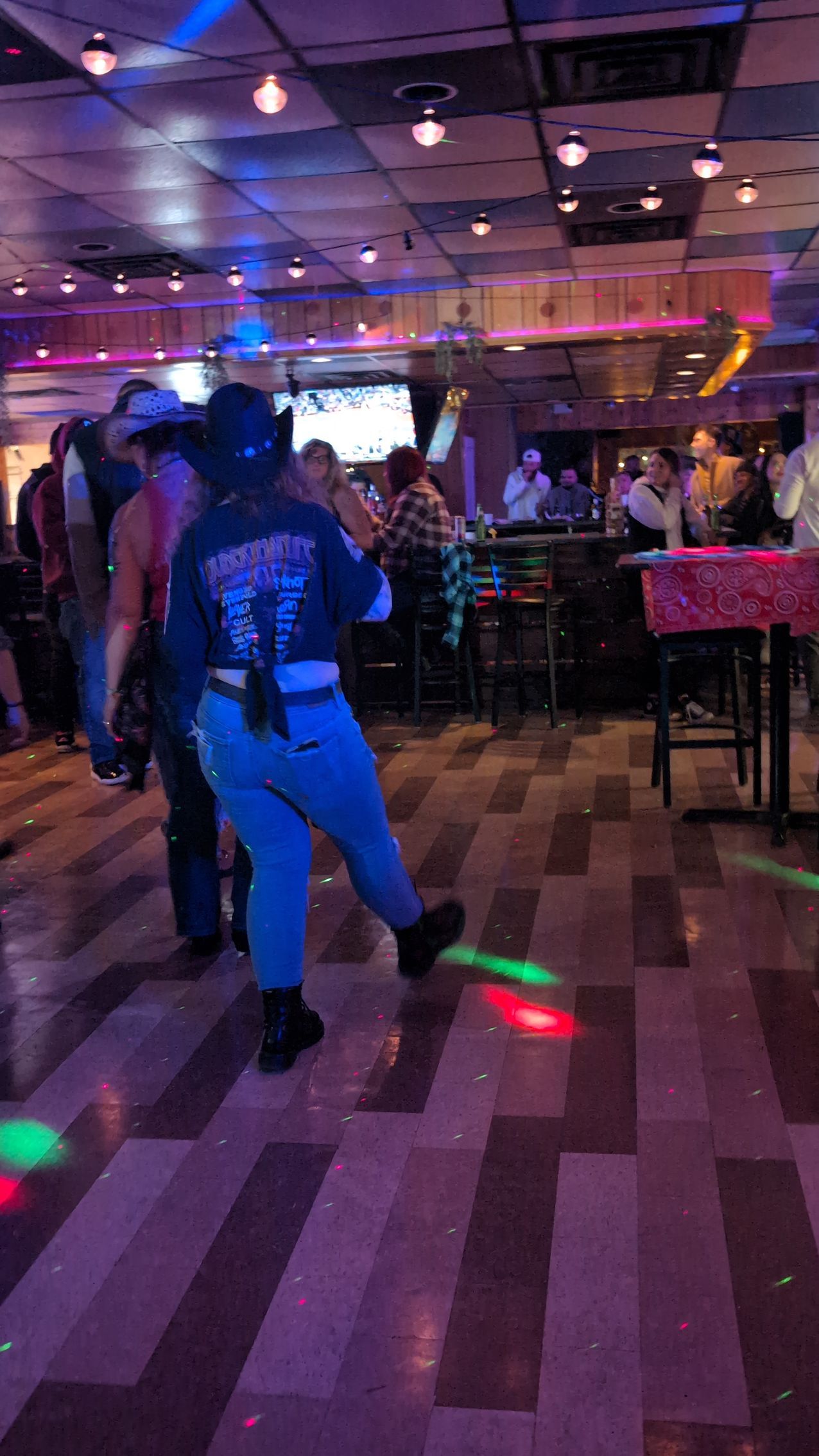 Dimly lit bar with people dancing near tables and colorful floor lights