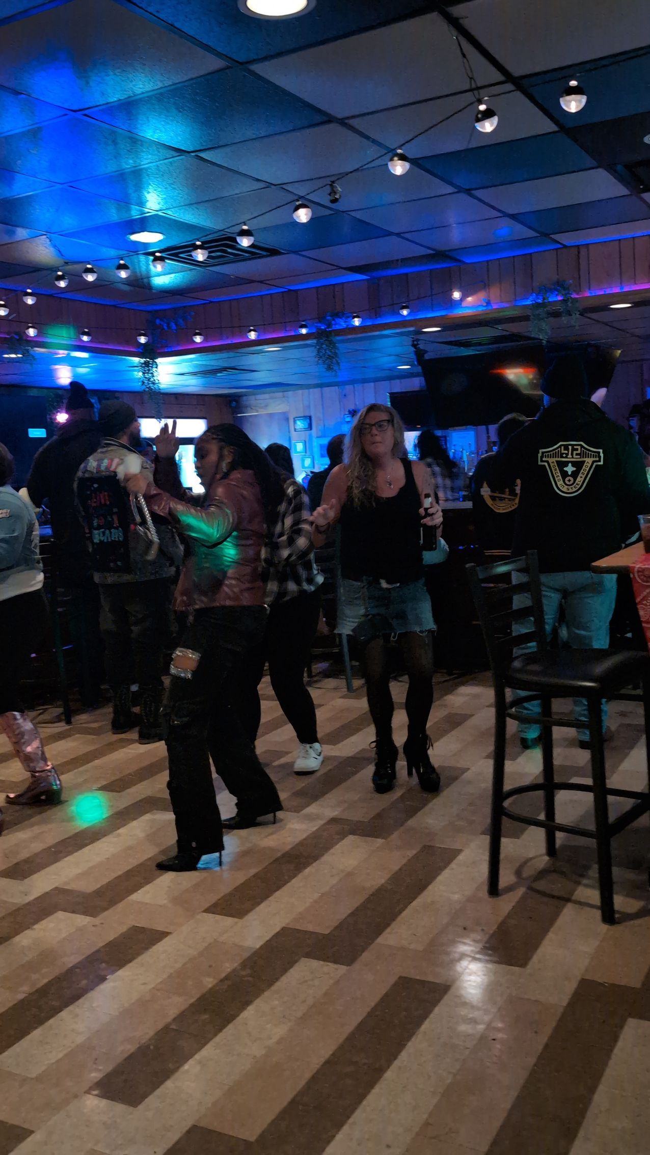 People standing and chatting in a dimly lit bar with blue ceiling lights and a checkered floor.