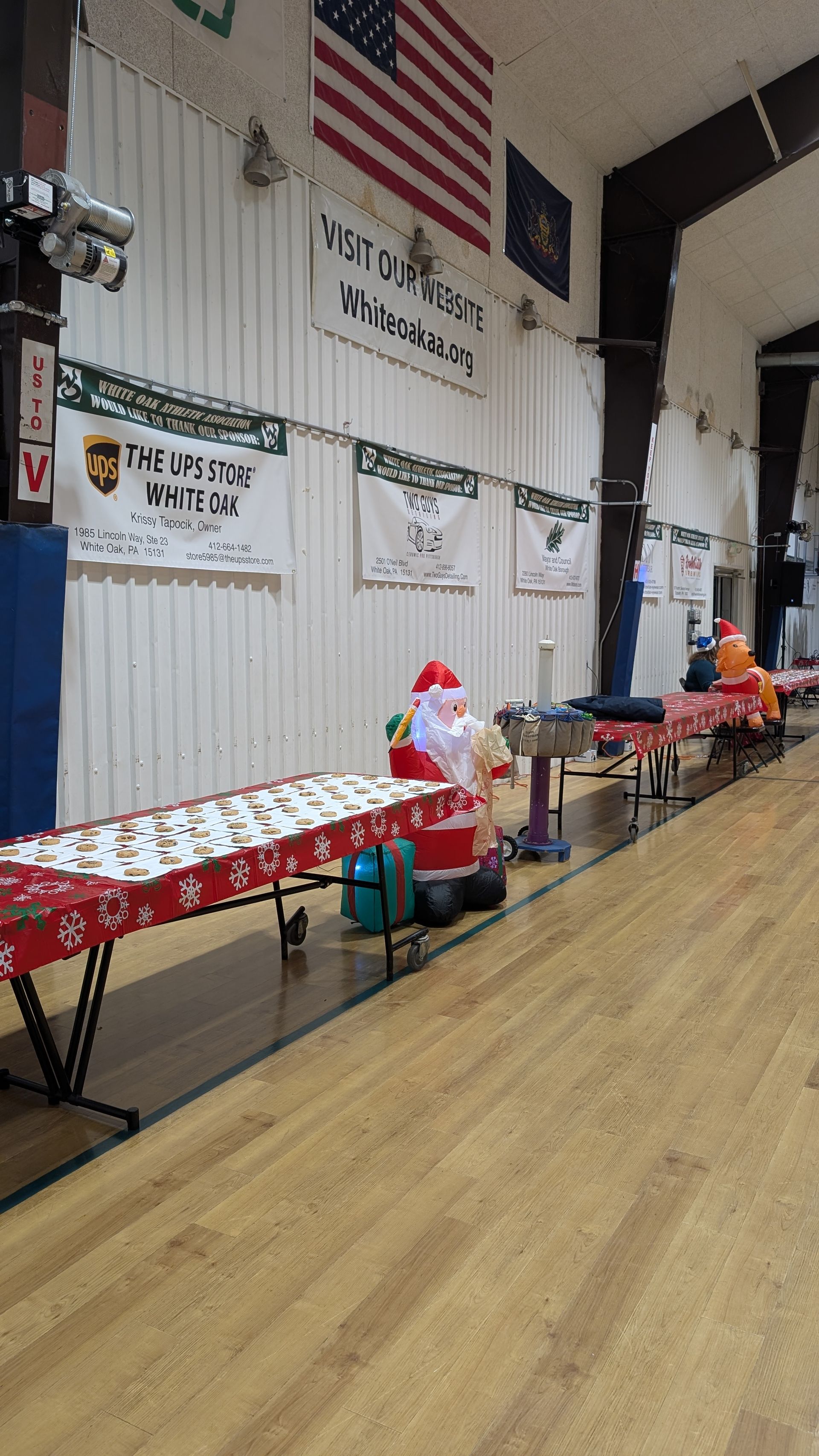Holiday display tables with Santa figurines and wrapped gifts in a gymnasium under American flags
