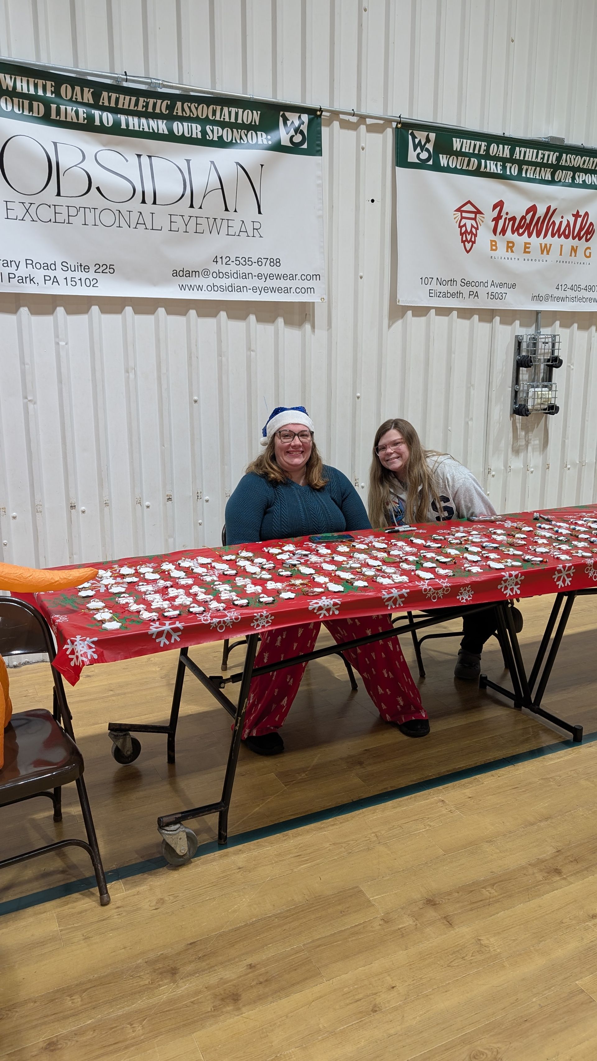 Two people seated behind a red-patterned table at an indoor booth display, with banners on the wall behind them.