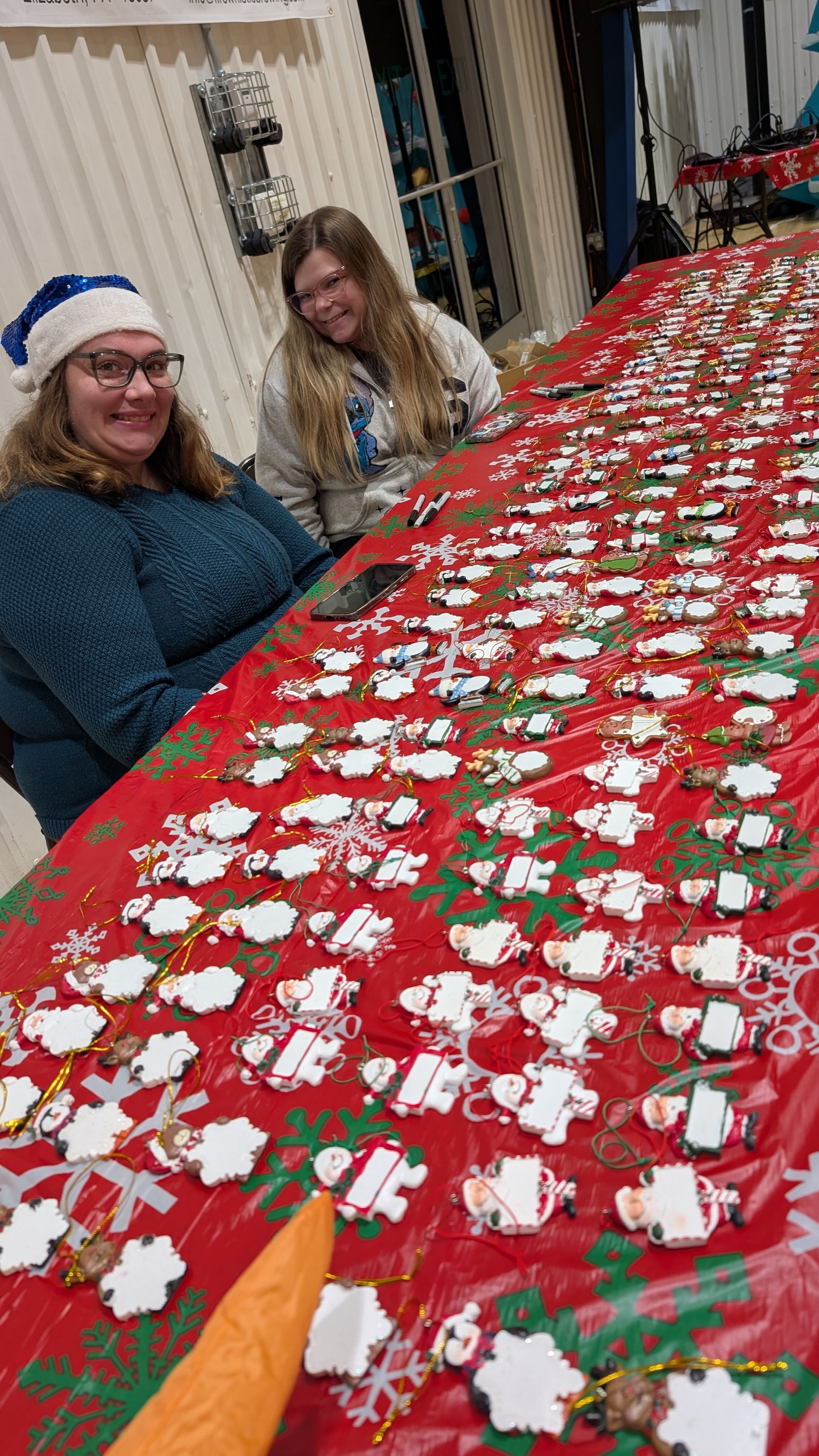 Two people sit beside a long table covered with red fabric and rows of small holiday crafts.