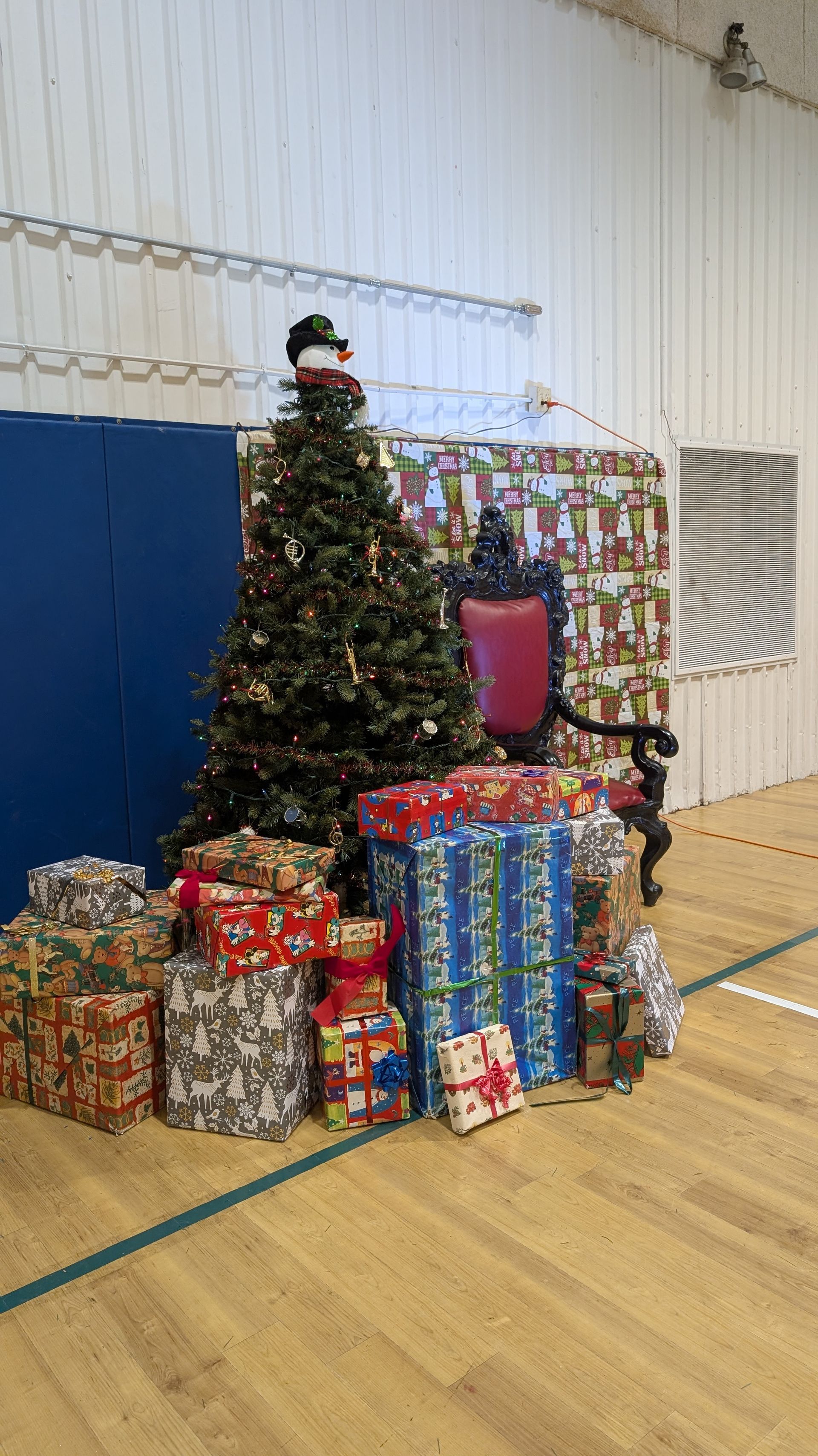 Decorated Christmas tree surrounded by wrapped gifts in a gymnasium with blue wall padding