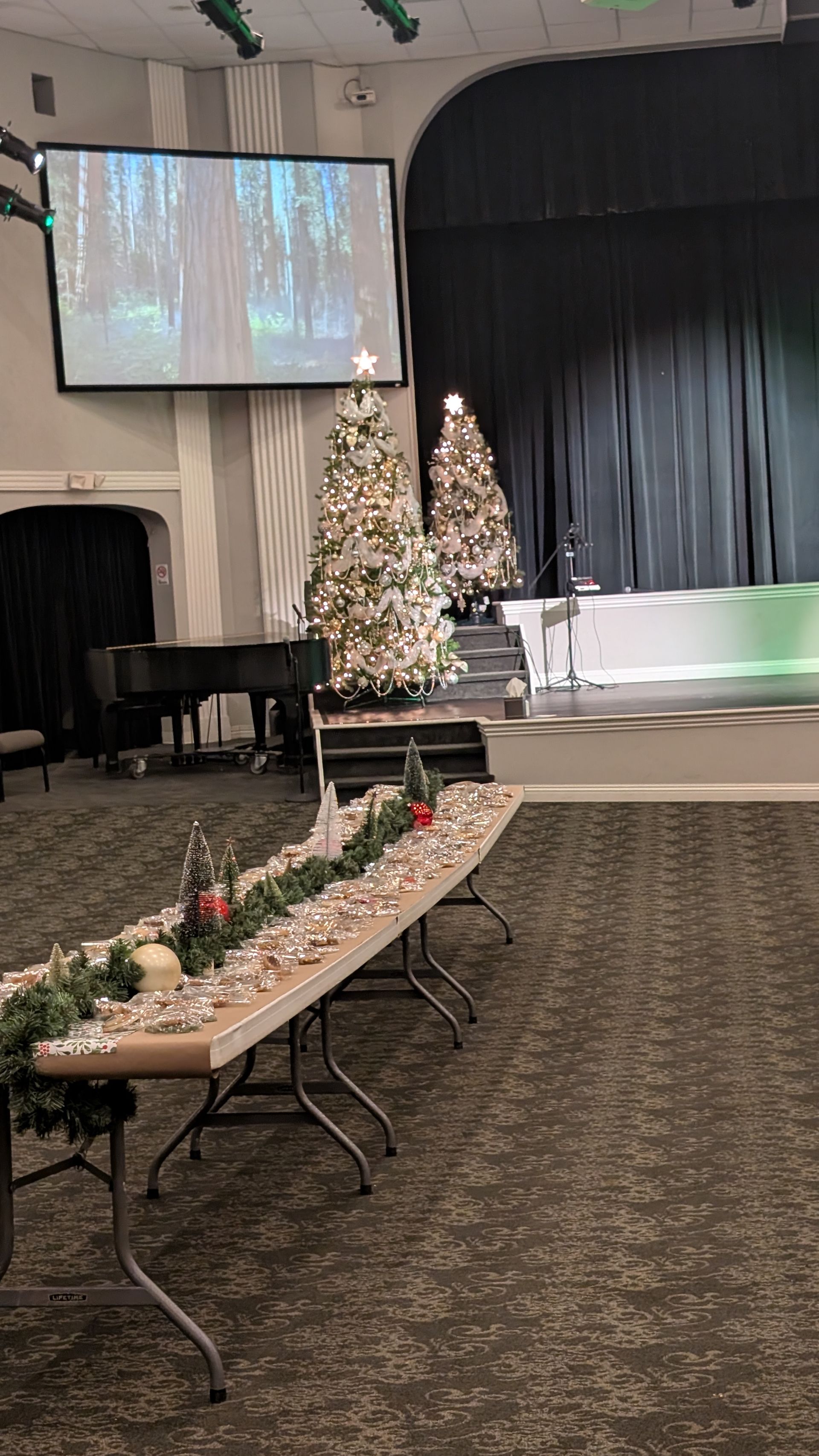 Indoor stage decorated with Christmas trees, floral table arrangements, and a projection screen in a banquet hall