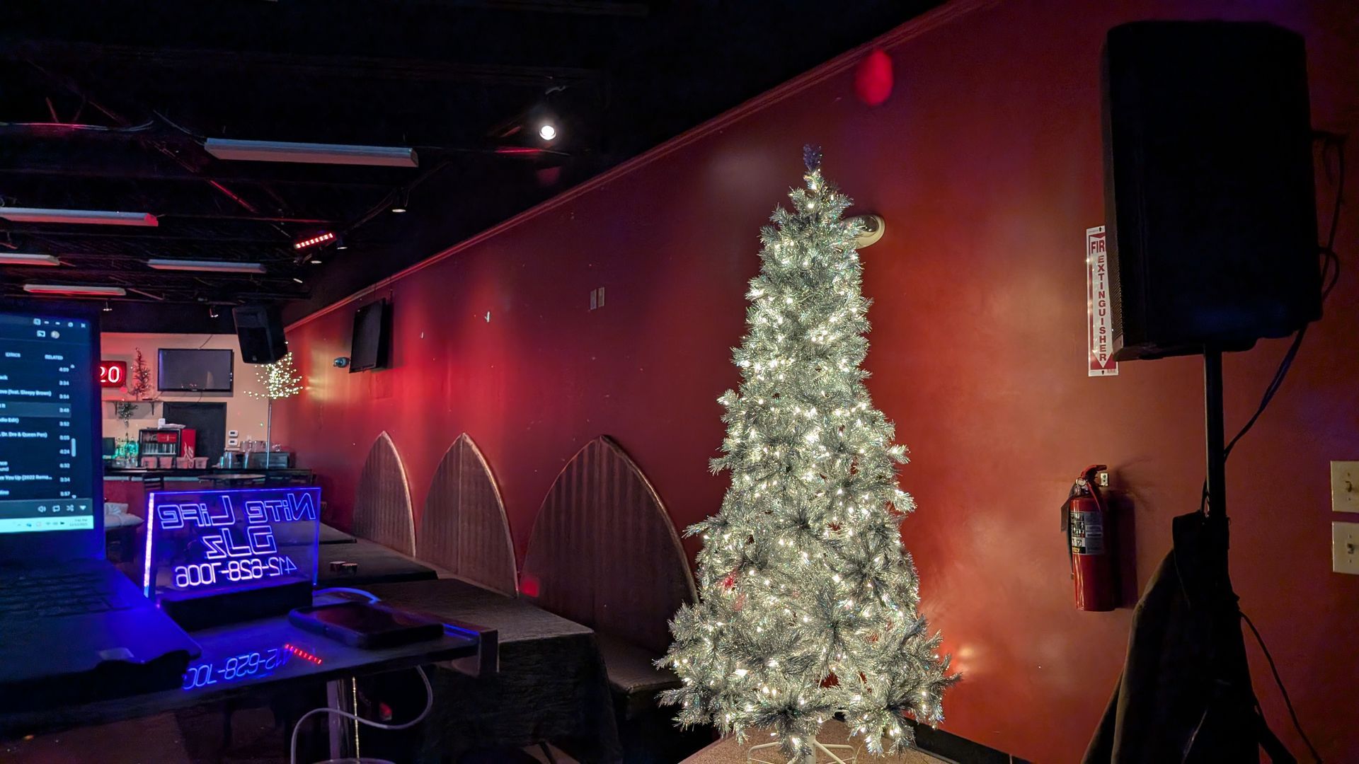 Dim lounge with a lit white Christmas tree, red wall lights, and neon glow from a bar area