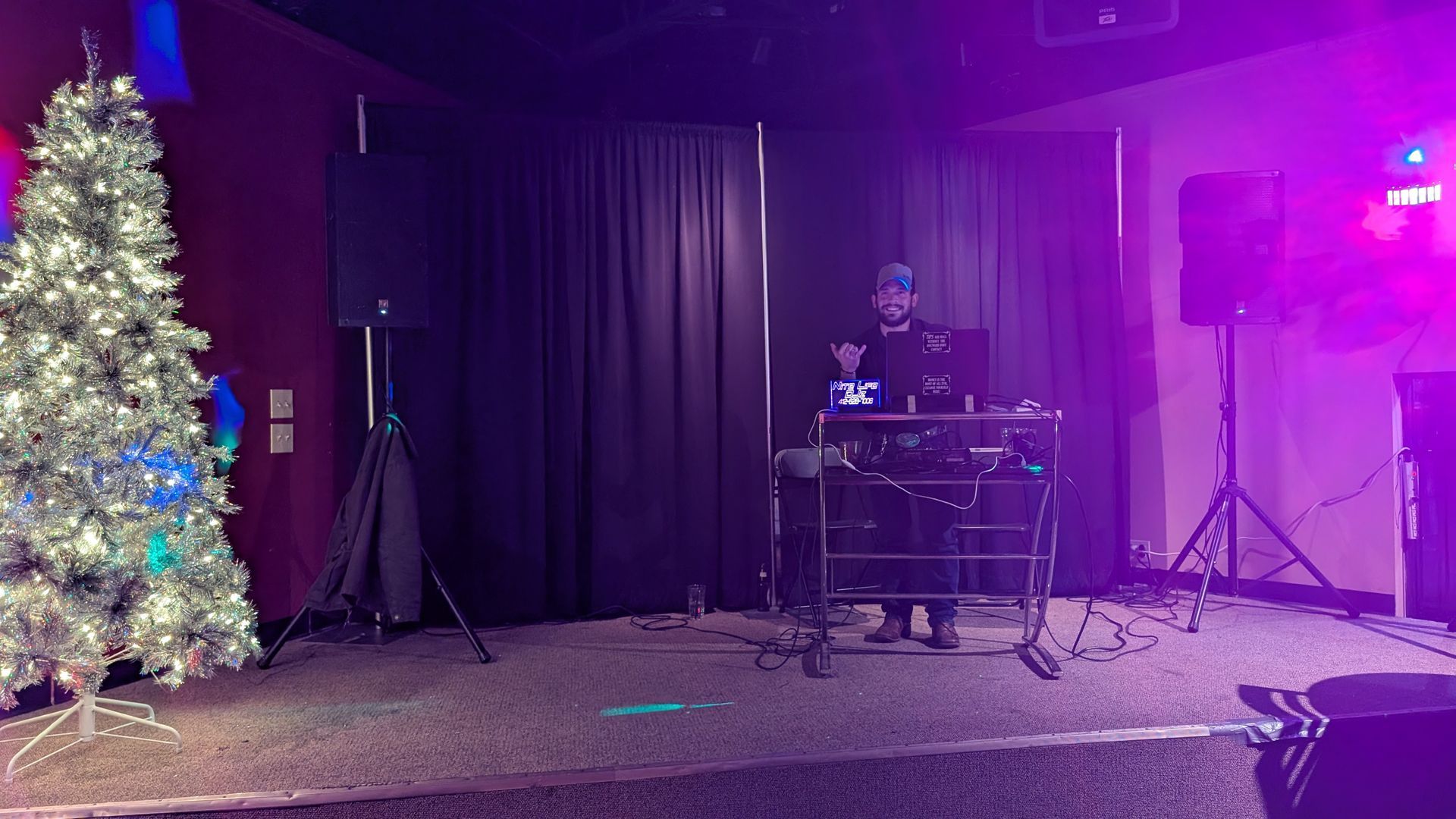 DJ performing on a small stage beside a lit Christmas tree with purple lights and black curtains