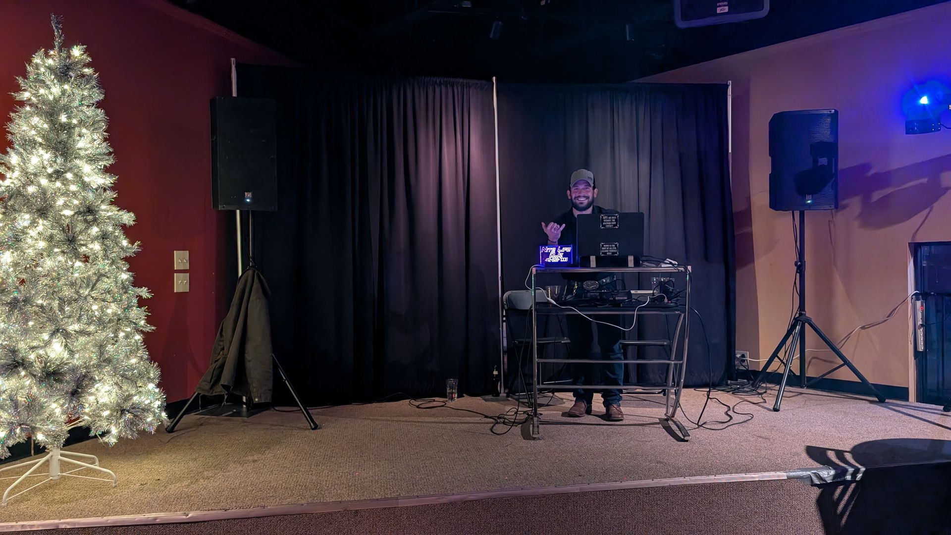 DJ setup on a small stage beside a lit Christmas tree, with black curtains and speakers in the background