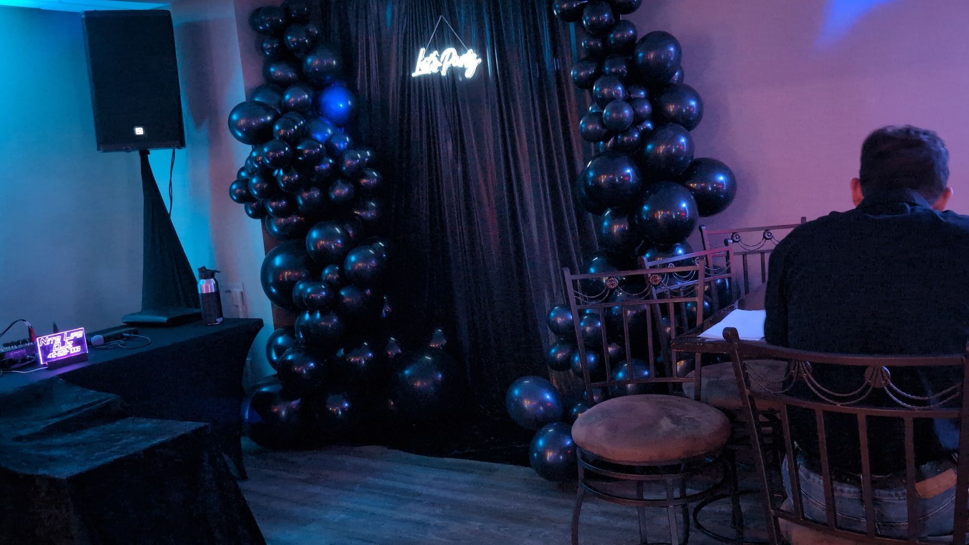 Dimly lit event space with black balloon arches framing a curtain, blue-purple lighting, and seated guests.
