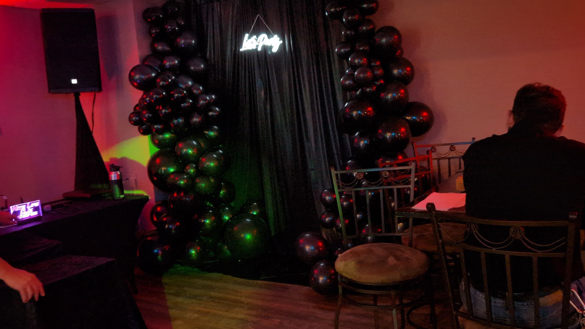 Dim lounge with black balloon arch, red-green lighting, and a seated person at a table