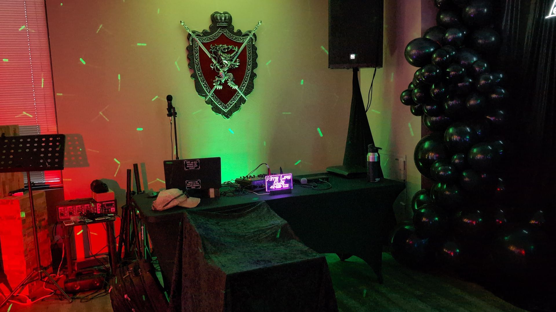 Dim room with red and green lighting, a desk and chair, wall crest, and black balloon arch on the right.