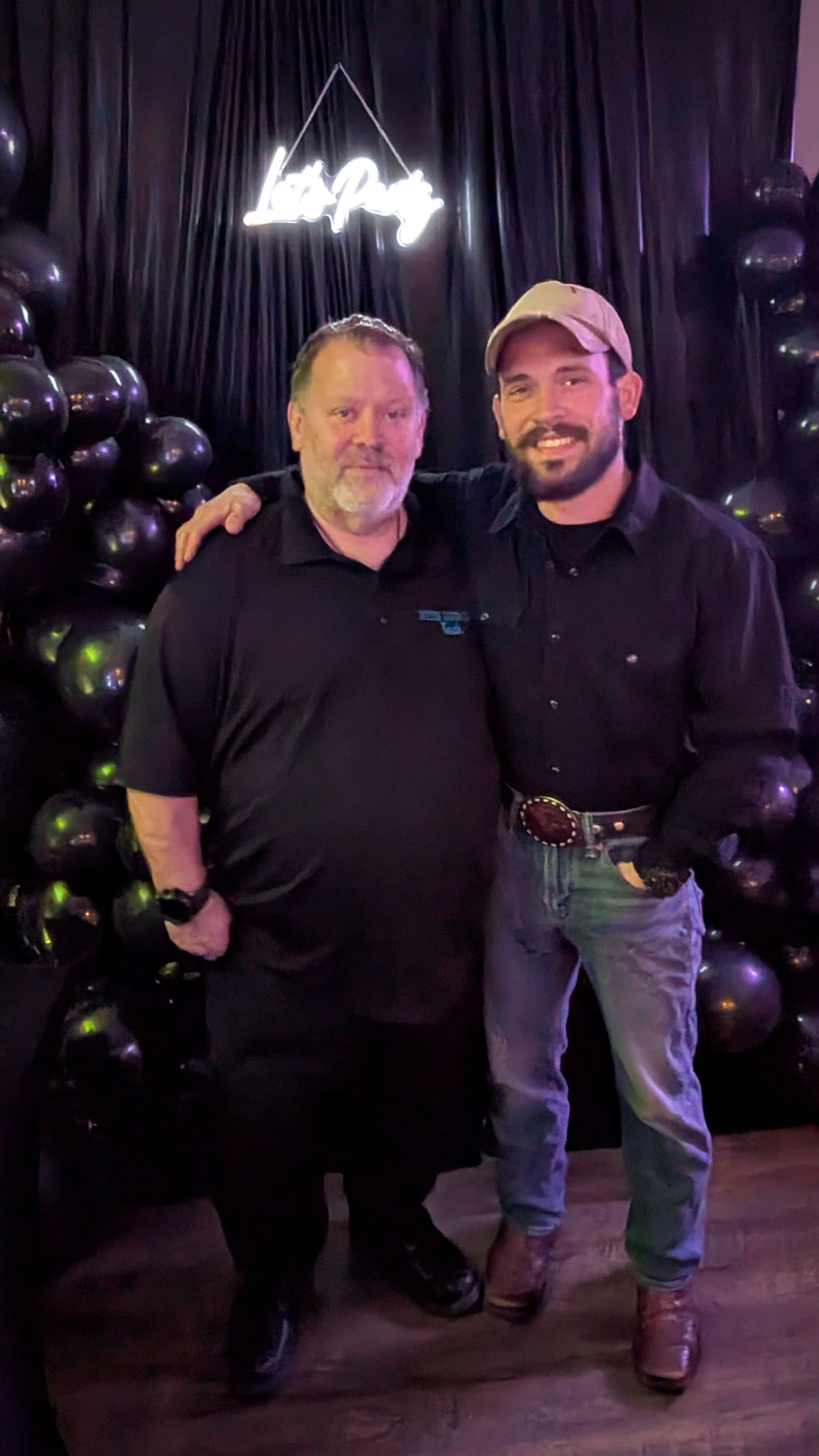 Two men pose together in front of black balloons and a neon sign at a party backdrop.