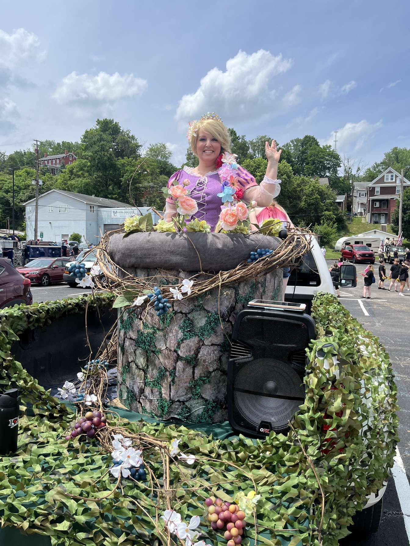 Person in a floral top waving from a flower-covered parade float outdoors under a blue sky