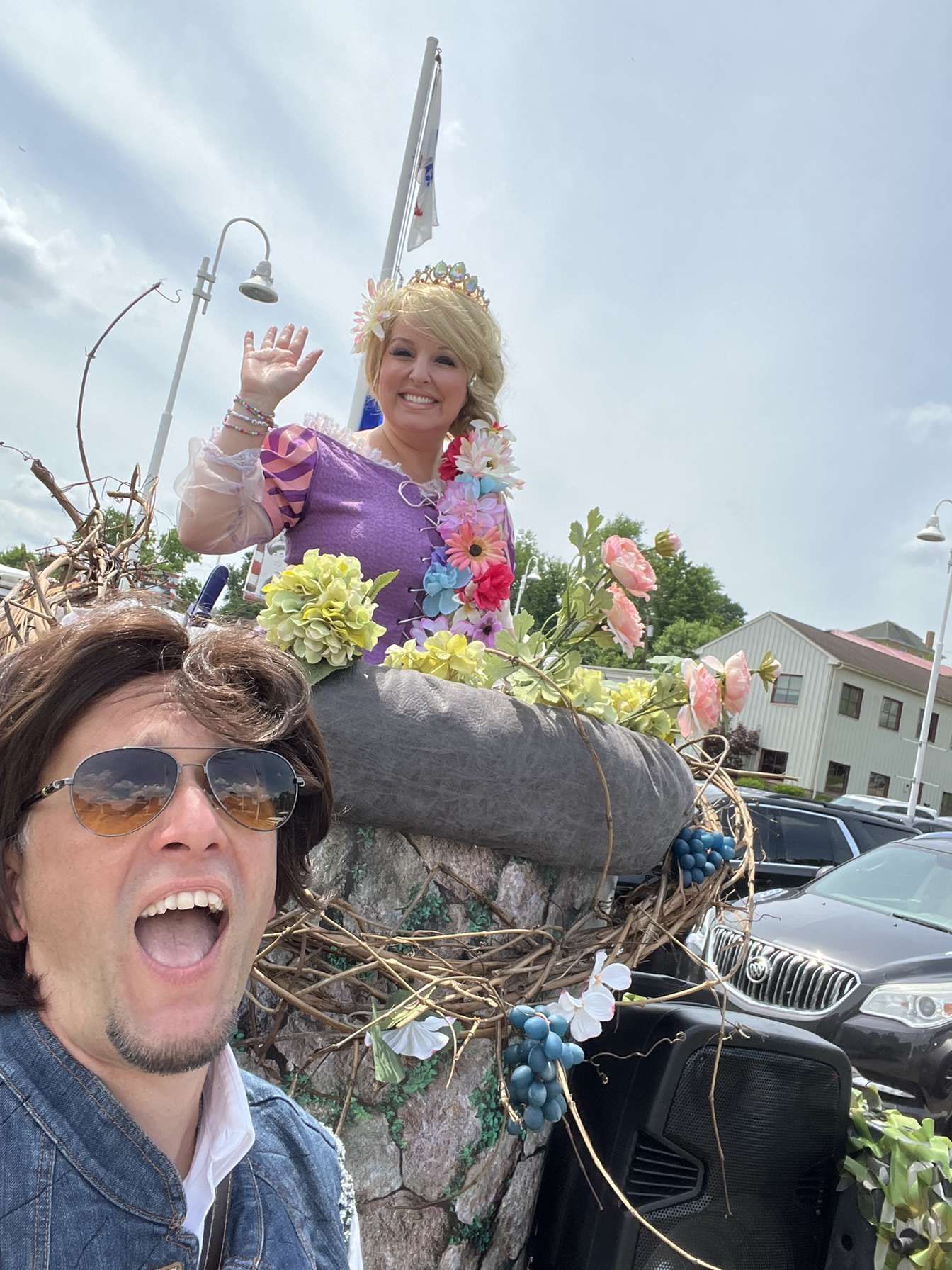 Smiling person in sunglasses posing with a woman in a purple dress on a flowered parade float outdoors