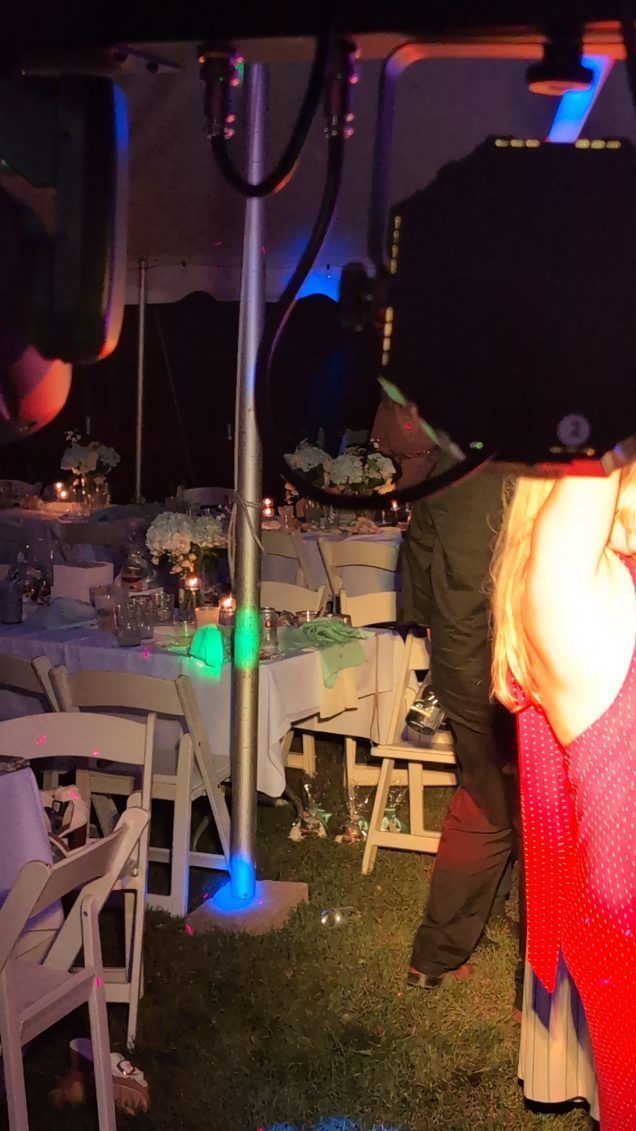 Dimly lit outdoor party with tables, chairs, and people near a white tent pole and colorful lights.