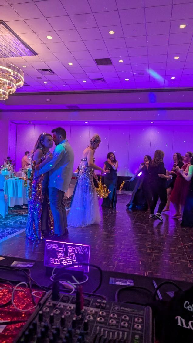 Couple slow dancing at a purple-lit indoor wedding reception, with guests gathered around.