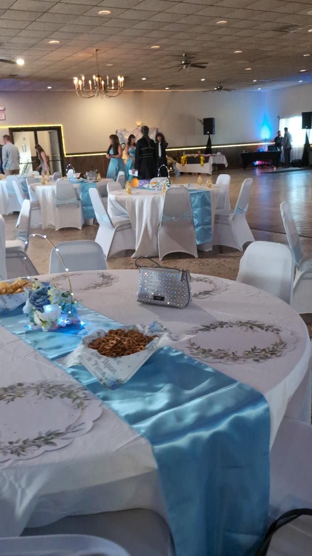 Decorated banquet hall with round tables, white chair covers, blue runners, and guests near the back