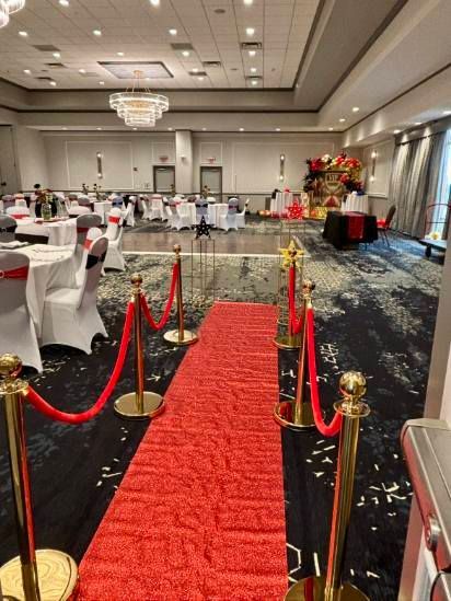 Banquet hall with red carpet aisle, gold stanchions, and round tables under chandeliers