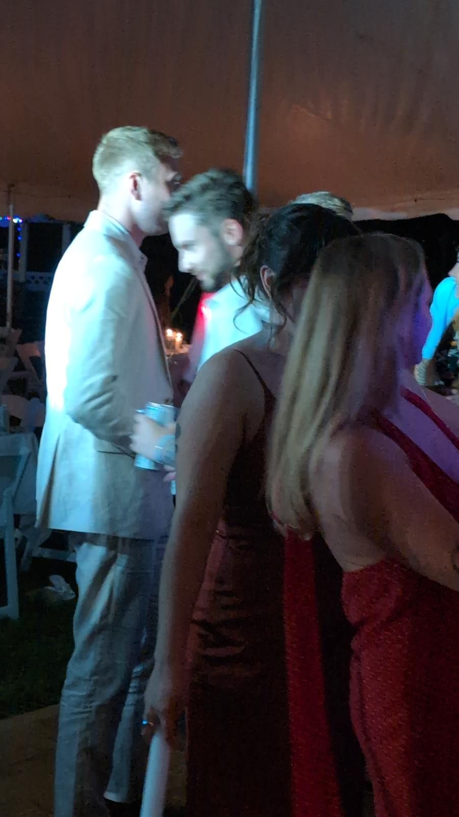 People mingling at a dimly lit party, with blue and pink lights and a wooden wall in the background