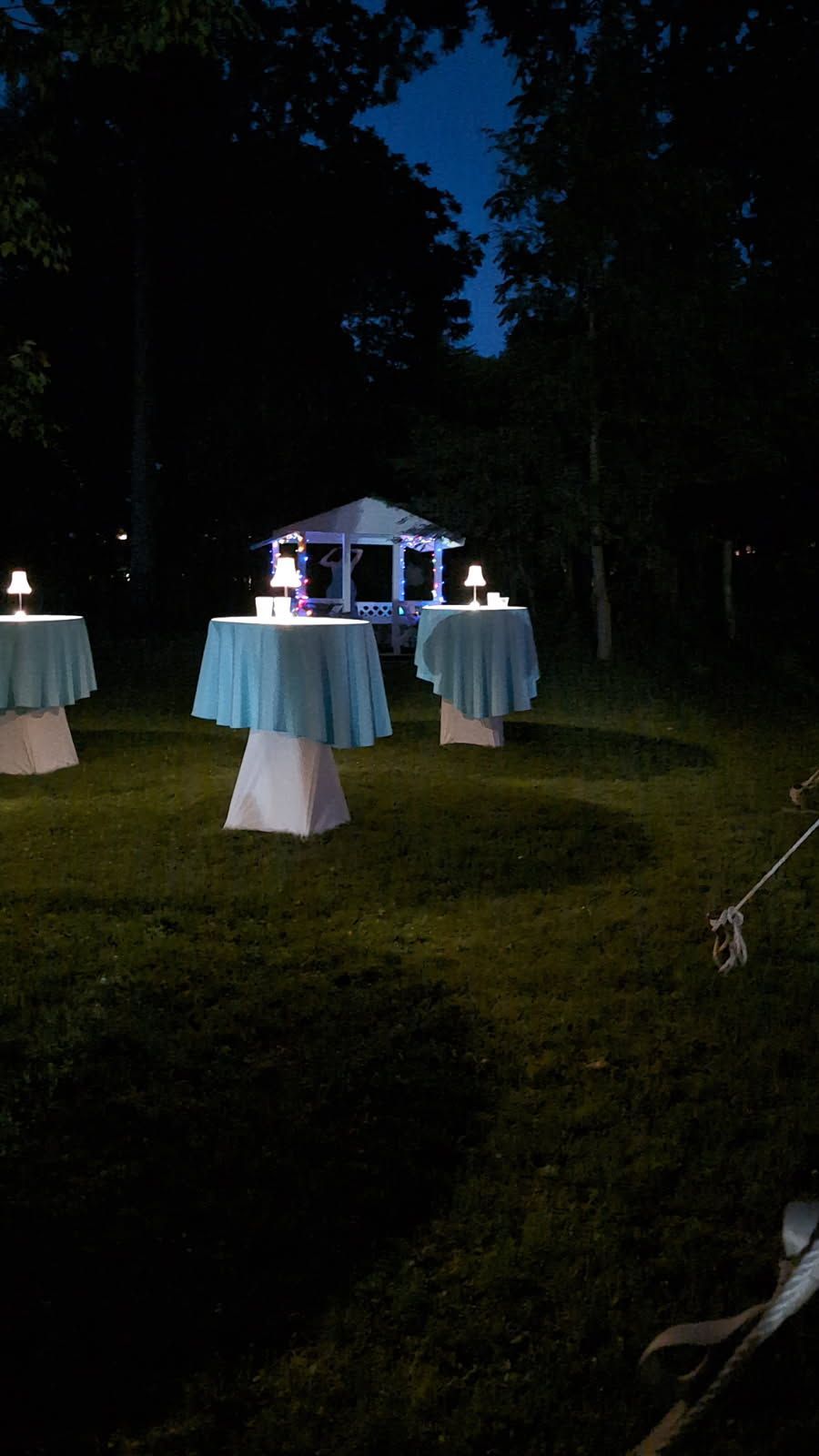 Nighttime outdoor reception with lit cocktail tables on a lawn and a glowing pavilion in the background