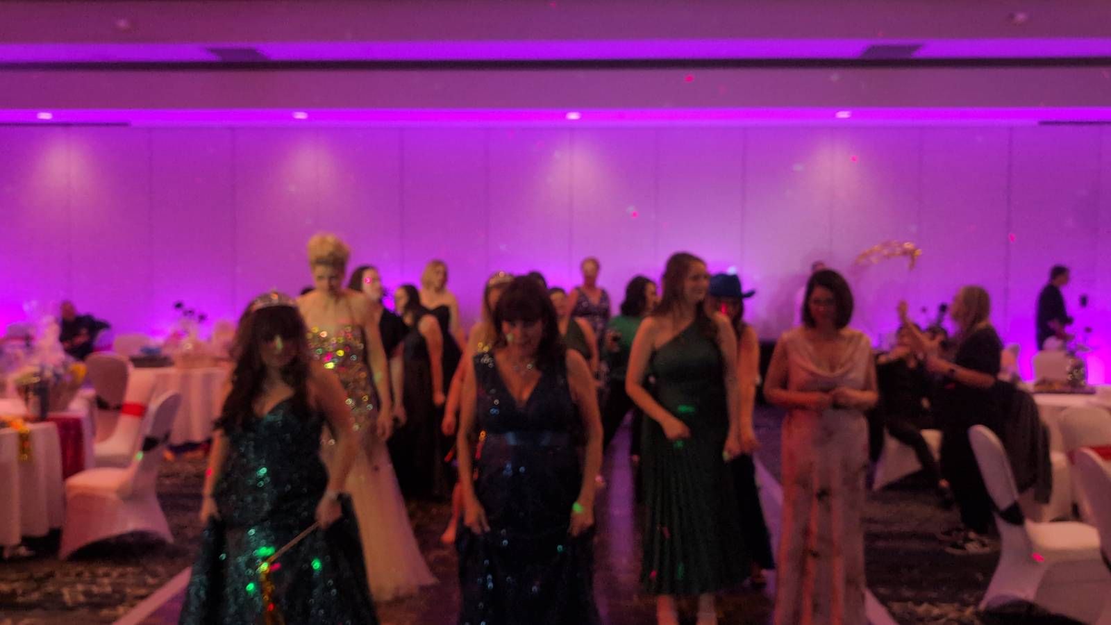 People in formal gowns walking through a purple-lit banquet hall during an event