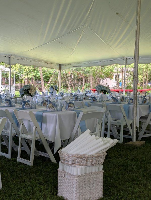 Round tables set for an outdoor event under a white tent, with white chairs and a basket of folded linens.