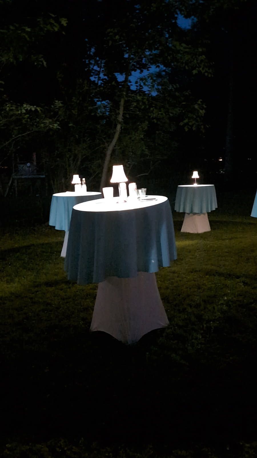 Round cocktail tables lit in a dark outdoor garden at night, with white tablecloths and glowing centerpieces
