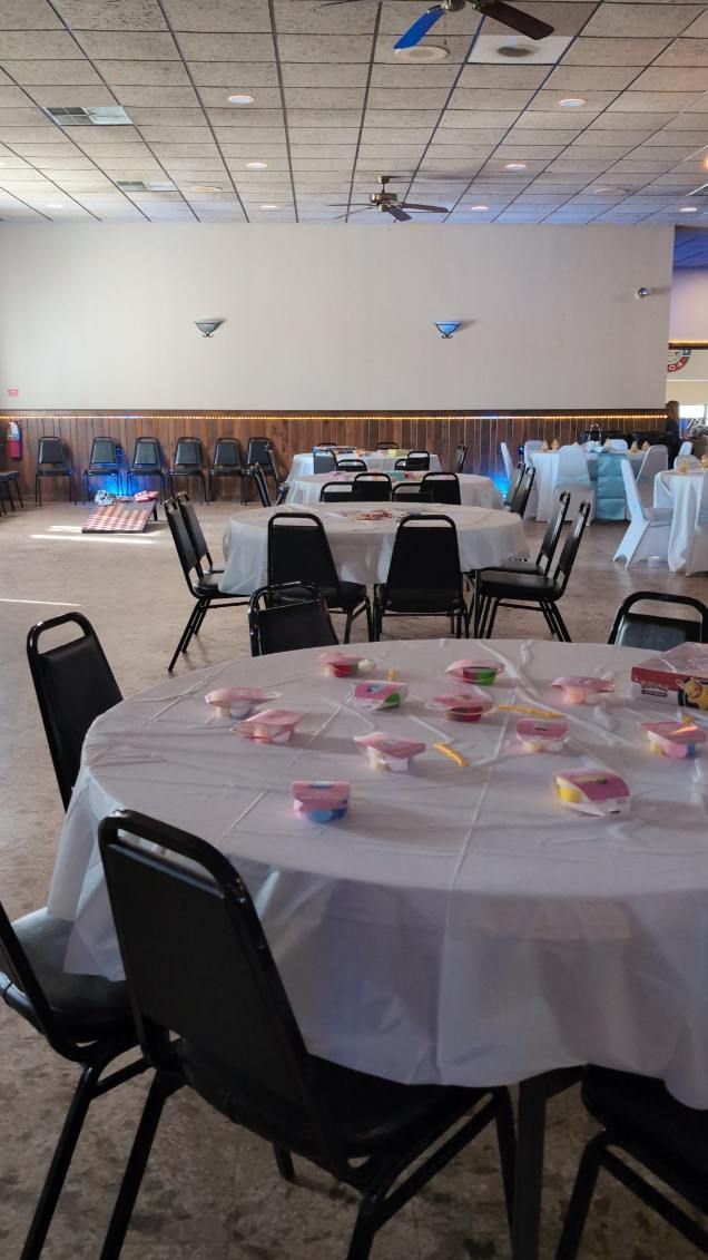 Round tables with white tablecloths and black chairs set up in a banquet hall with tiled ceiling and wall lights.