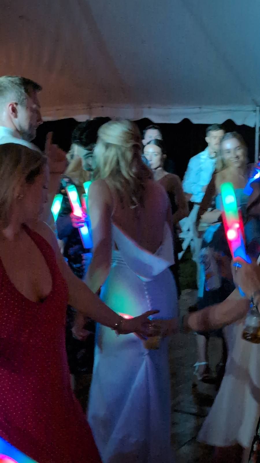 People dancing at a lively party under a tent, holding glowing neon light sticks.