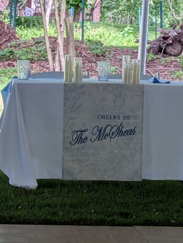 Outdoor Mother’s Day table with white cloth and candles, reading “Cheers to The Mothers”