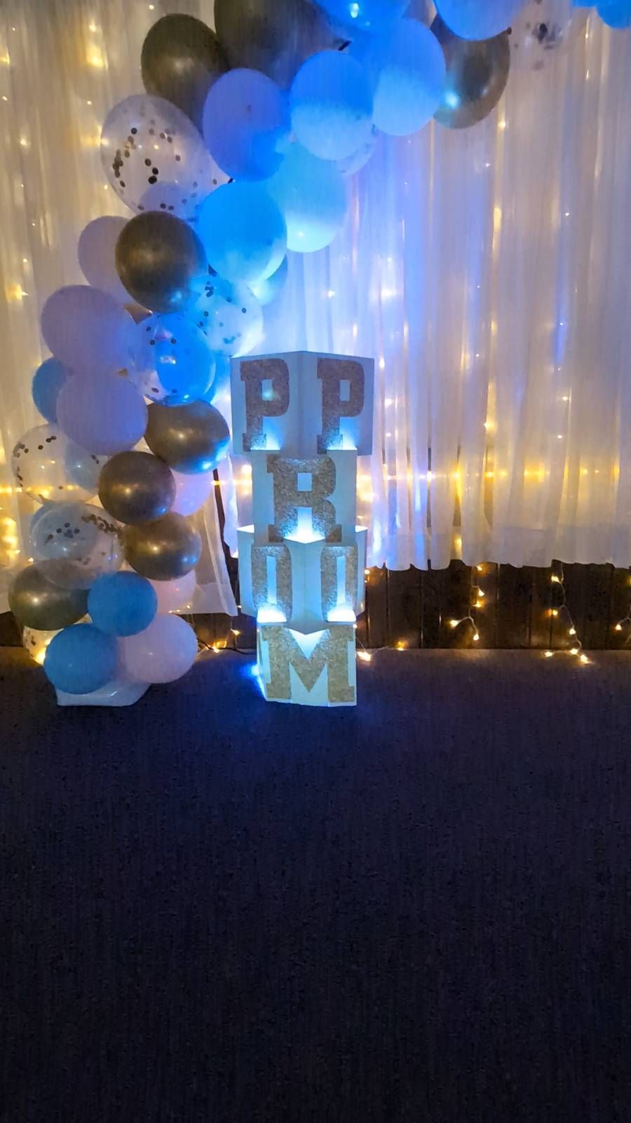 Blue and silver balloon backdrop with glowing “HAPPY” letters and warm string lights on a dark floor