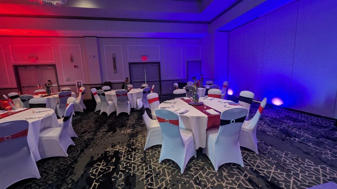 Red-and-blue lit banquet hall with round tables, white chair covers, and scattered guests.