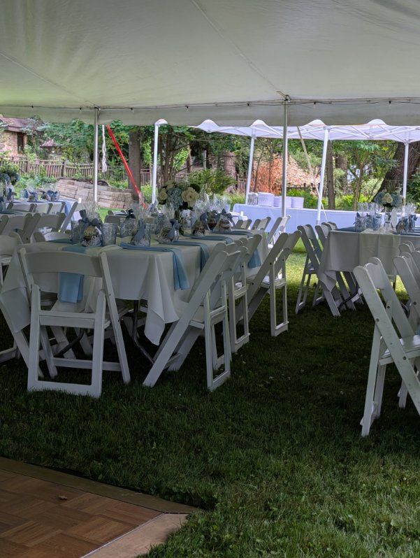 White folding chairs and tables set under a tent at an outdoor event on a grassy lawn.